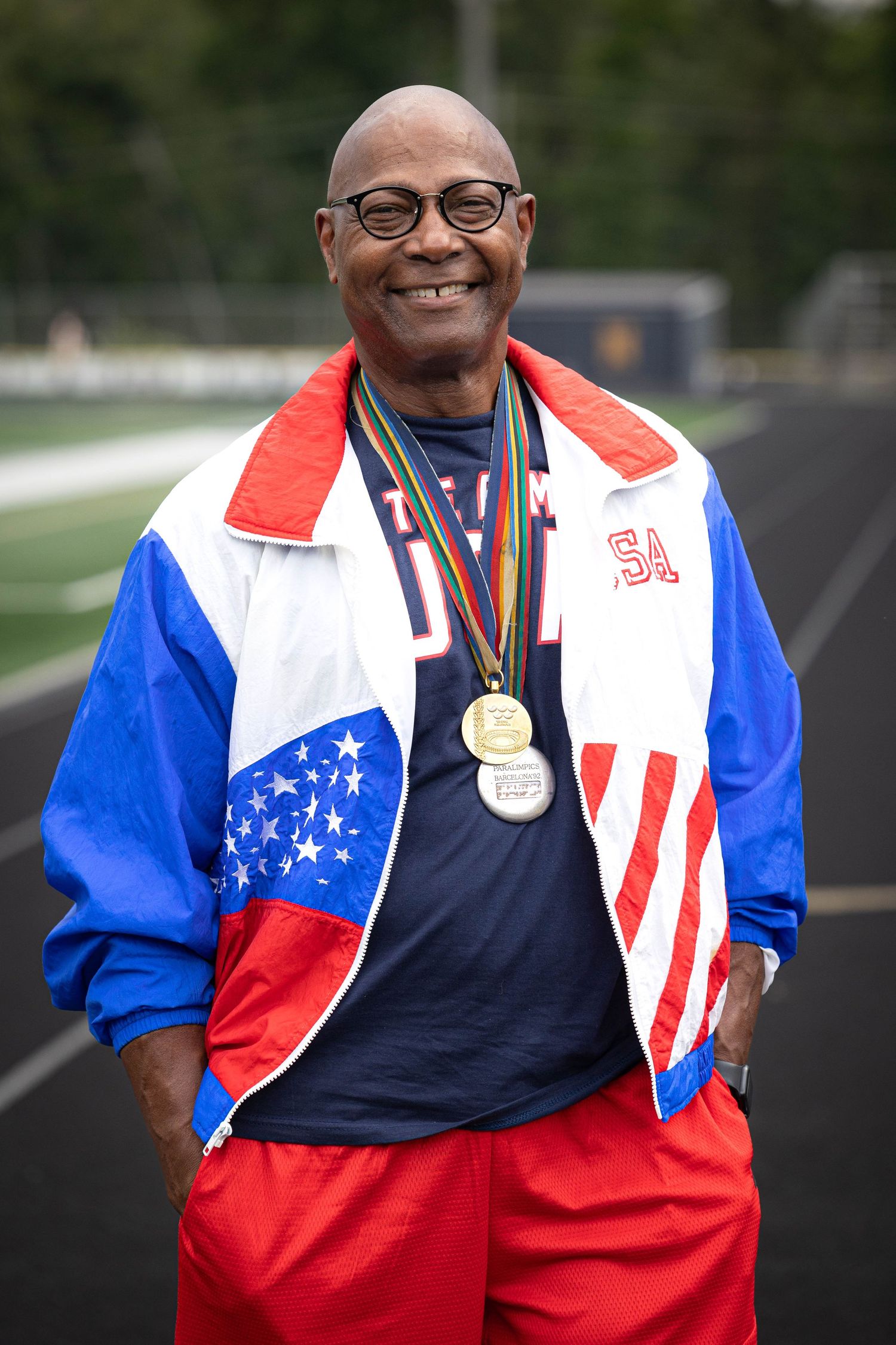 Athlete in patriotic jacket displaying medals against outdoor track background.