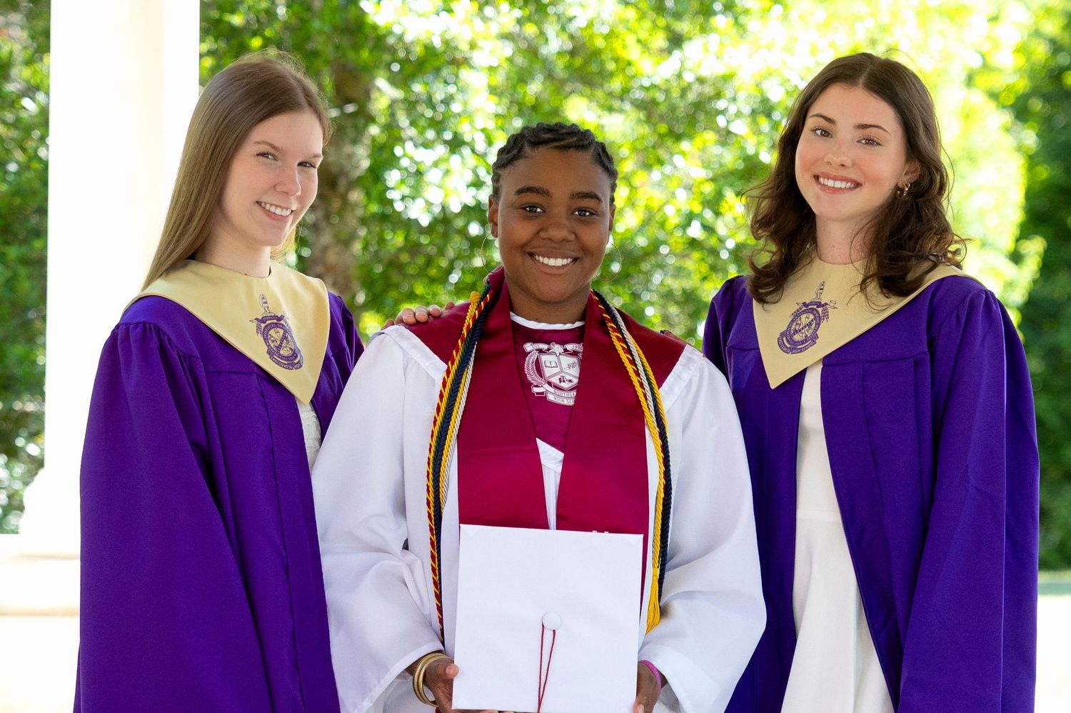 Three graduates in purple robes smile together at an outdoor graduation ceremony.