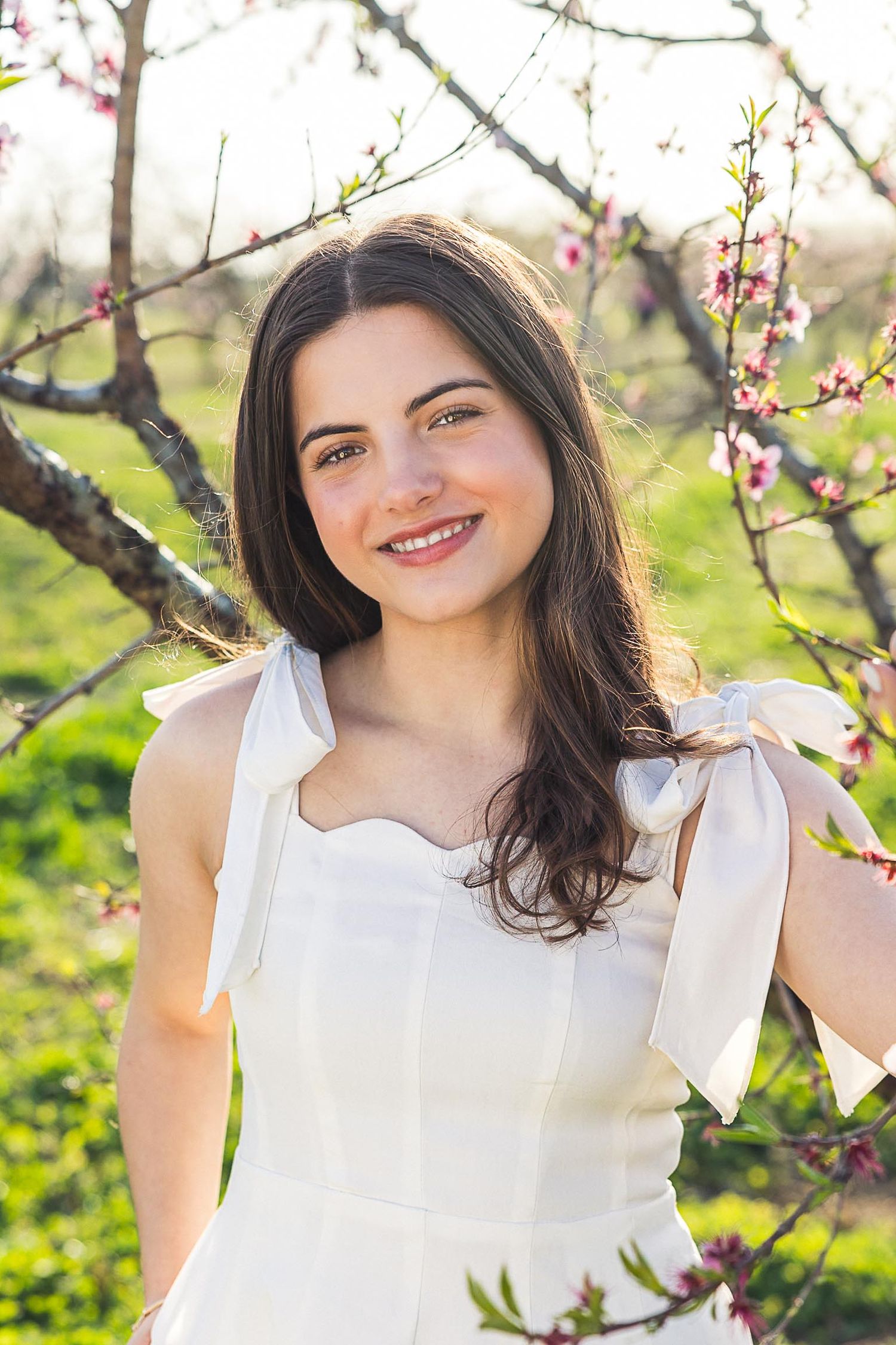 A person in a white dress smiles while standing among blooming spring trees in warm sunlight.