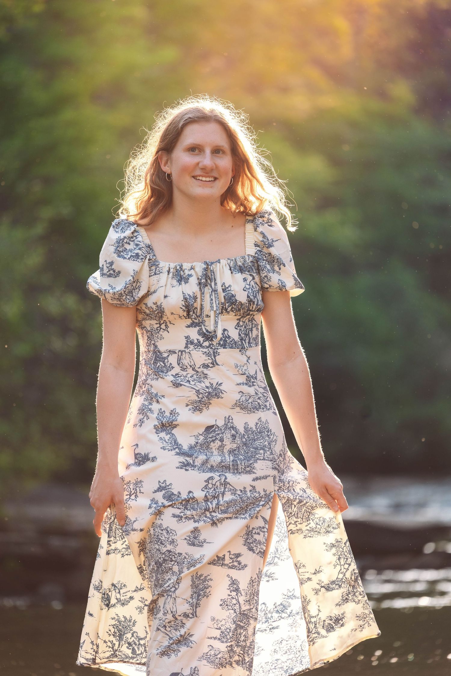 A person in a blue and white toile print dress poses by a stream during golden hour lighting.
