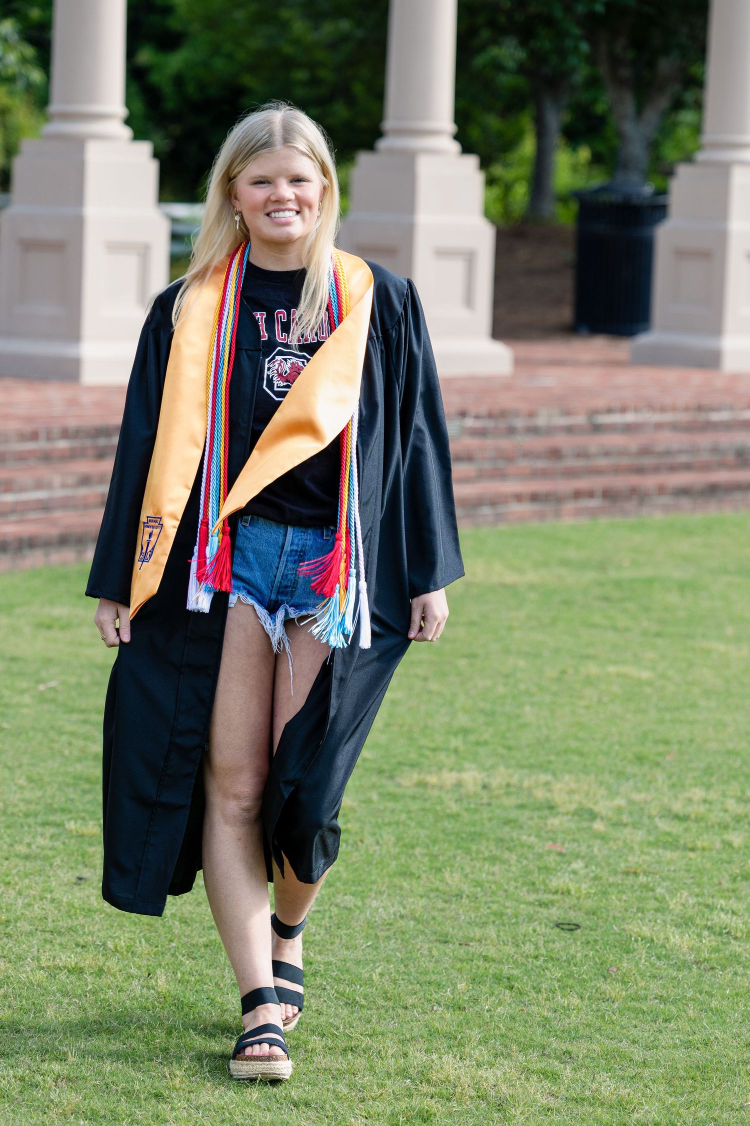 Graduate in black cap and gown with orange stole walks across green lawn near columned building.