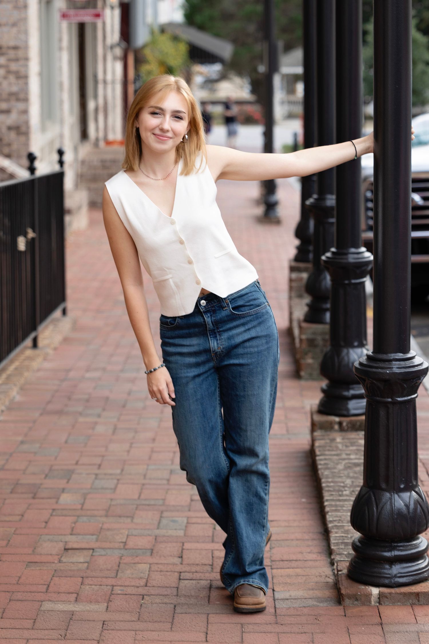 A person in blue jeans and white top poses playfully against black lamp posts on a brick sidewalk.