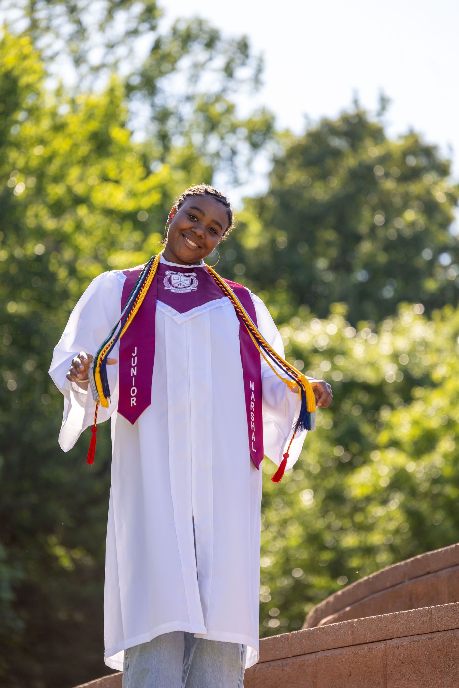 Smiling graduate in white cap and gown proudly displays colorful honor cords and sashes outdoors.