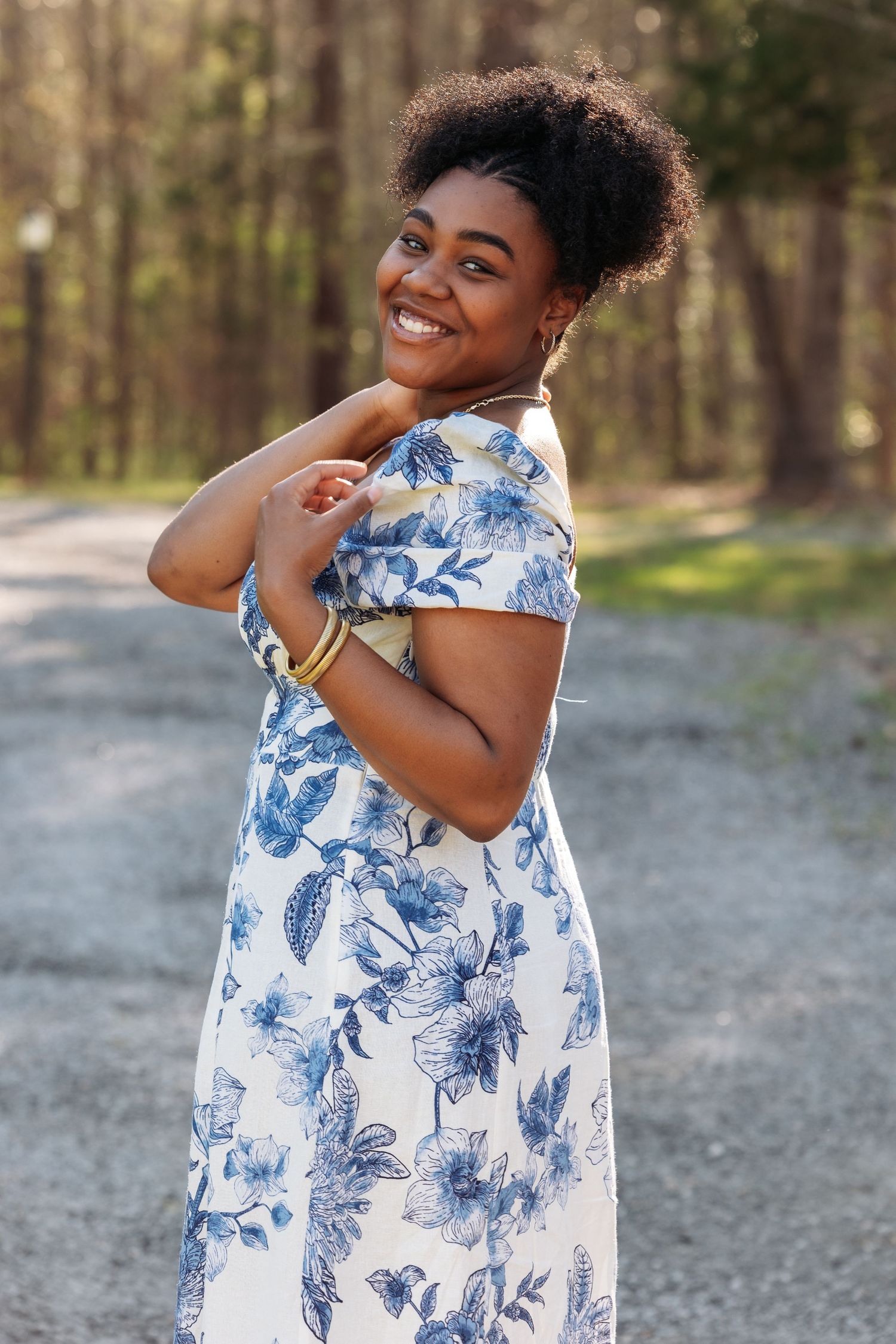 Smiling young Black woman in blue floral dress posing outdoors on a gravel path surrounded by trees.