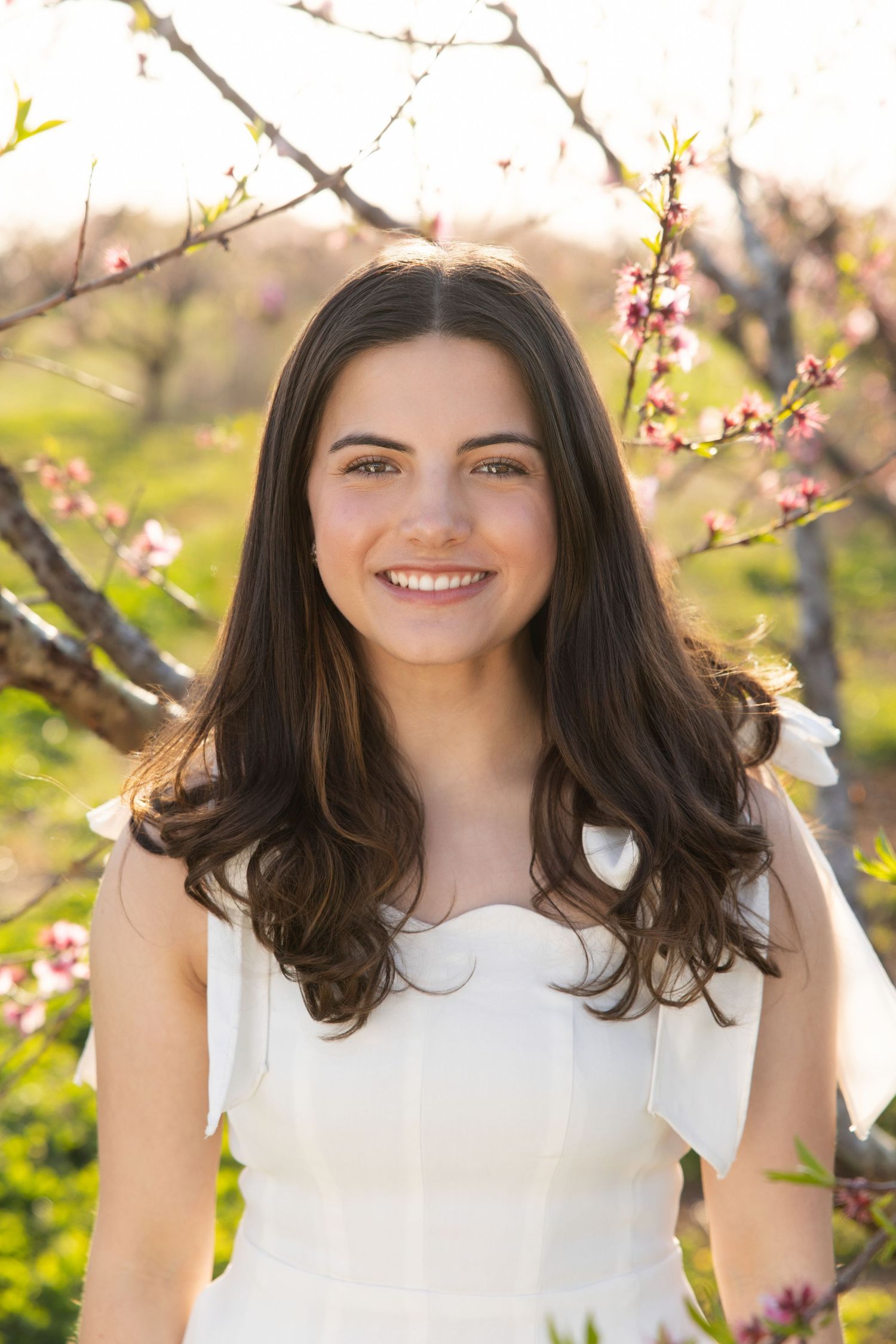 Portrait of someone with long dark hair smiling outdoors with blooming spring cherry blossoms in the background.