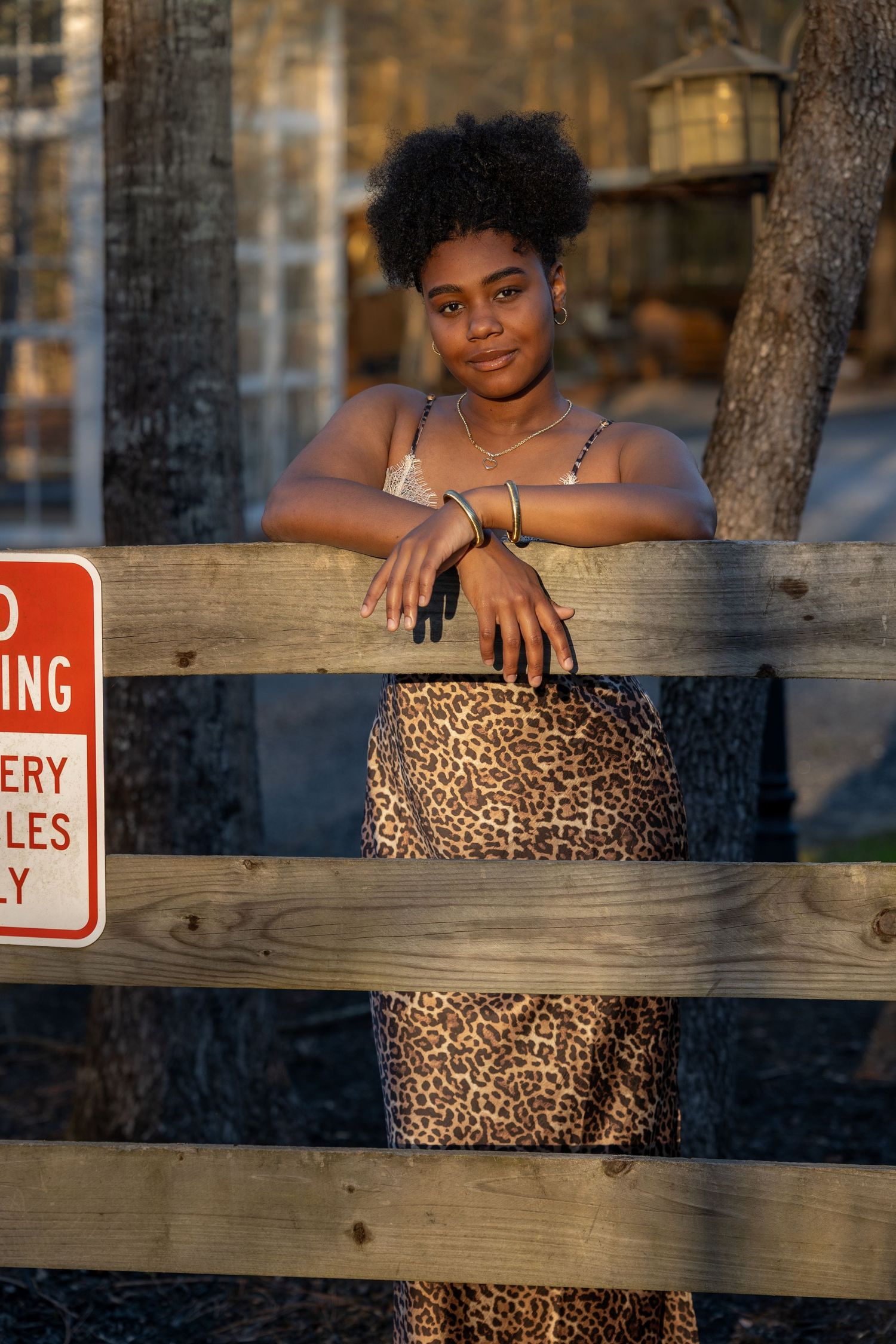 Young woman in leopard print skirt leans on wooden fence outdoors, wearing silver jewelry in warm golden light.