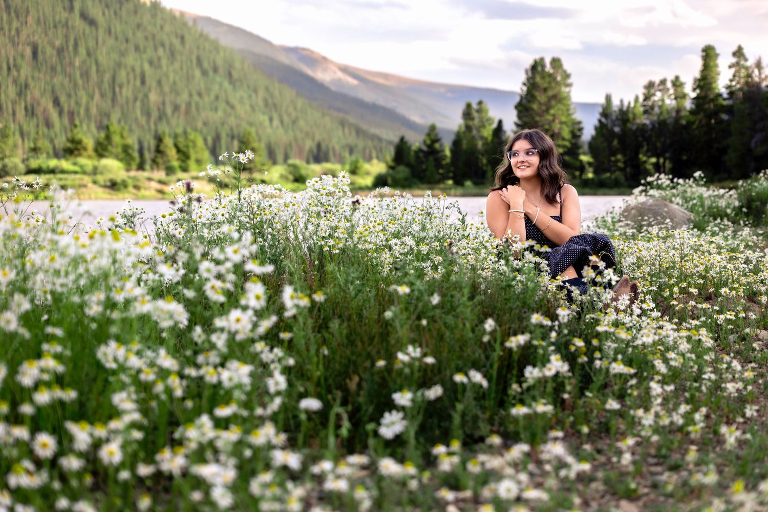 Young woman sitting in a field of white wildflowers with mountain forest backdrop at golden hour.