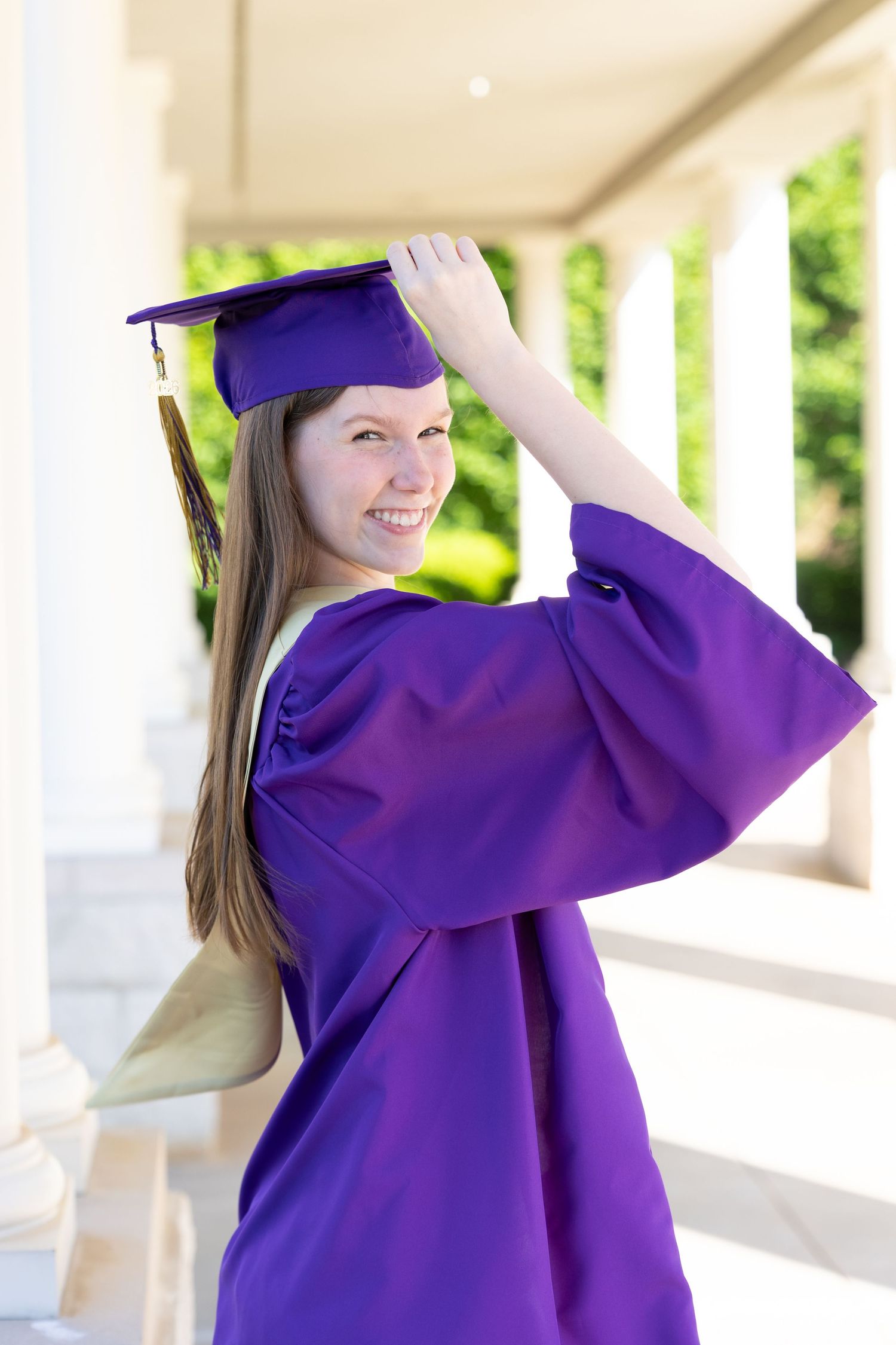 Smiling graduate in purple cap and gown poses outdoors between white columns on graduation day.