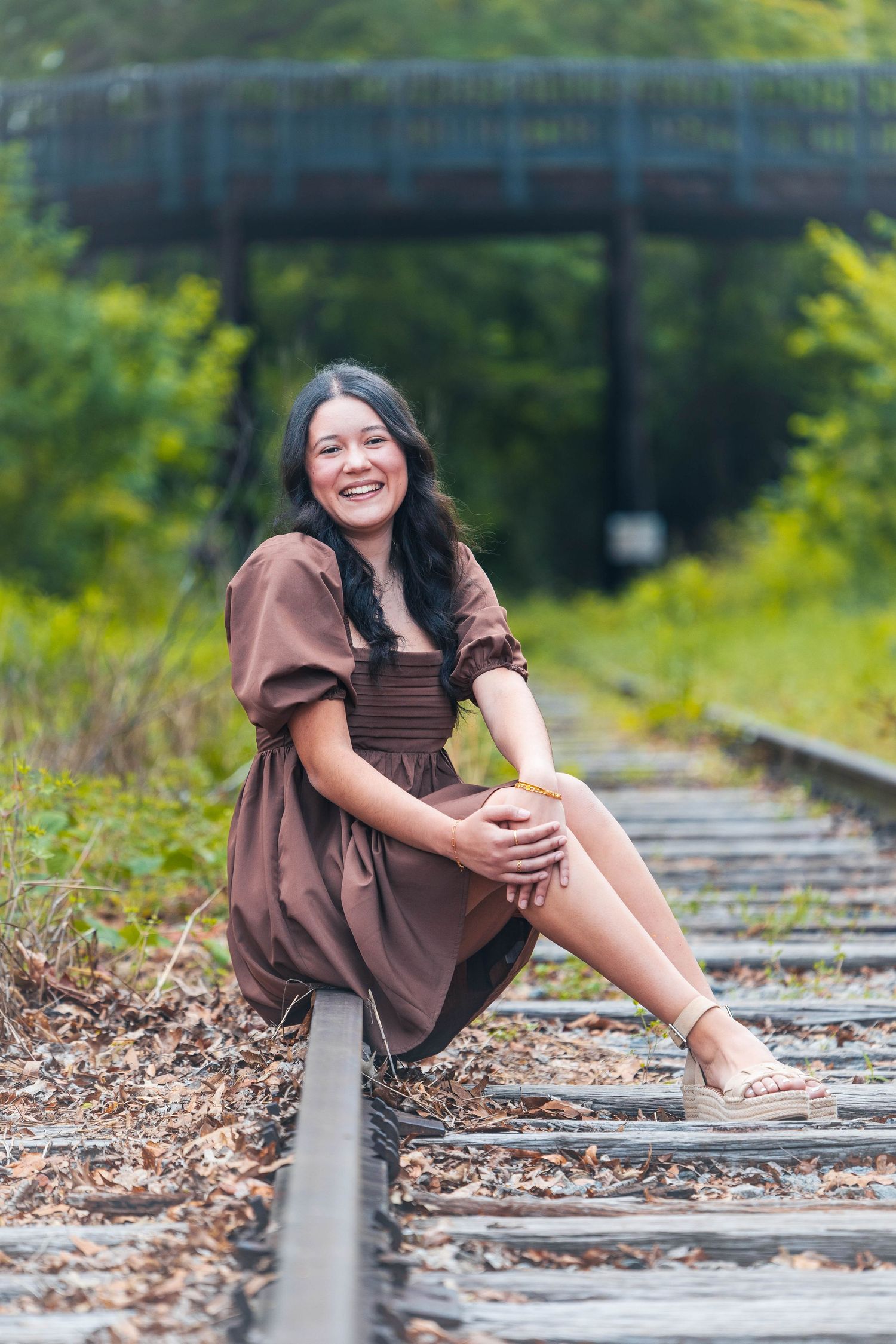 A person in a brown dress poses playfully on train tracks surrounded by lush greenery and fallen leaves.