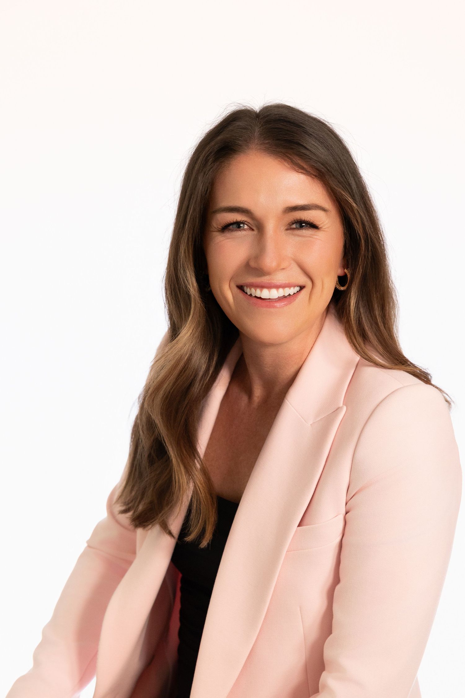 Smiling woman with brown hair wearing a pink blazer poses for a professional headshot on white background.