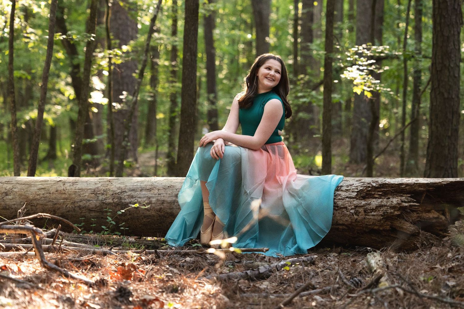Young woman in teal and pink gown sitting on a fallen log in a sunlit forest setting.