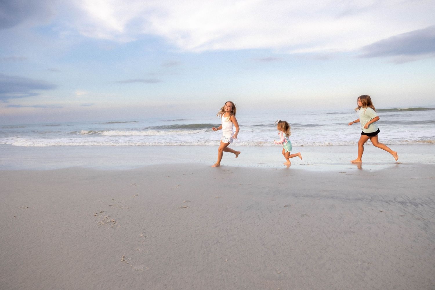 People running and playing together on a sandy beach with waves and cloudy sky in the background.