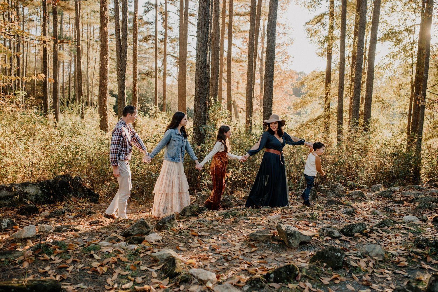 Wedding party members walk through sunlit autumn forest during golden hour photography session.