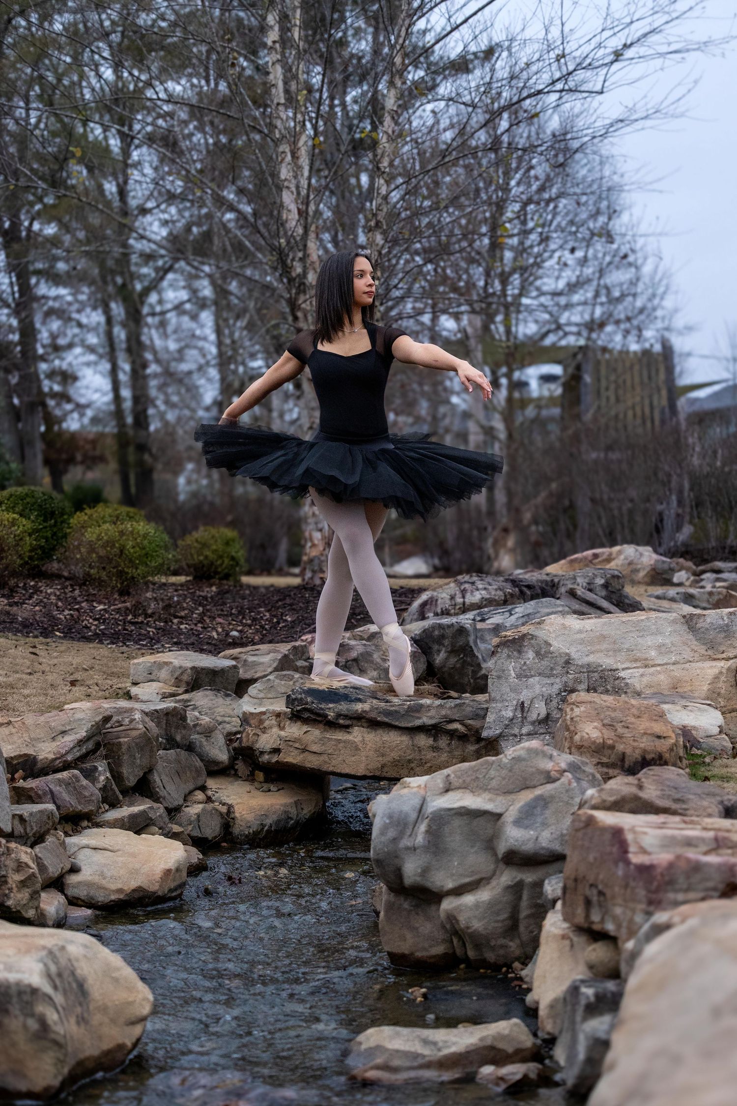 Dancer in black leotard and gray tights performs graceful poses on rocky stream bank in natural outdoor setting.