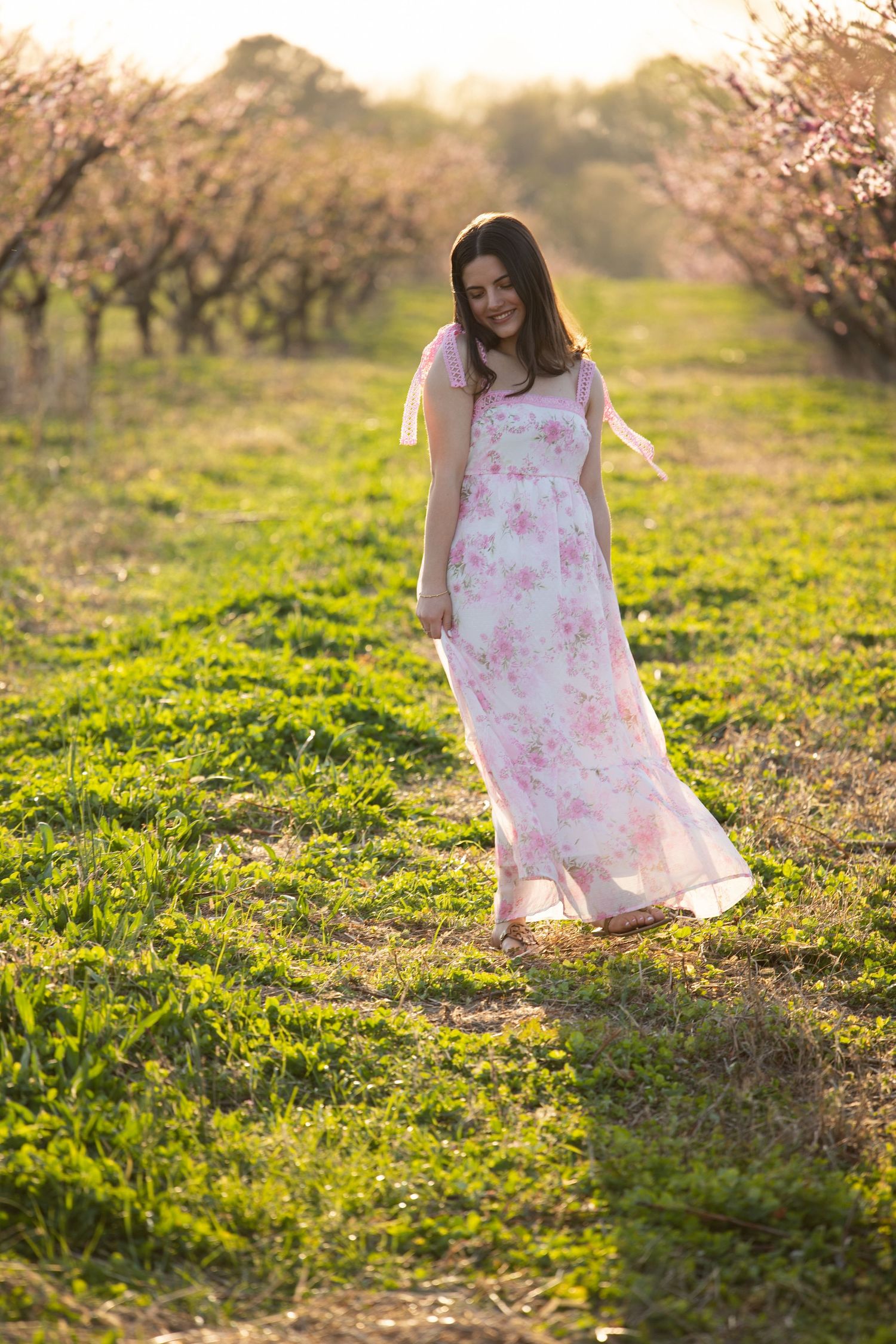 A person in a pink floral dress twirls joyfully in a sunlit meadow with blooming trees at golden hour.