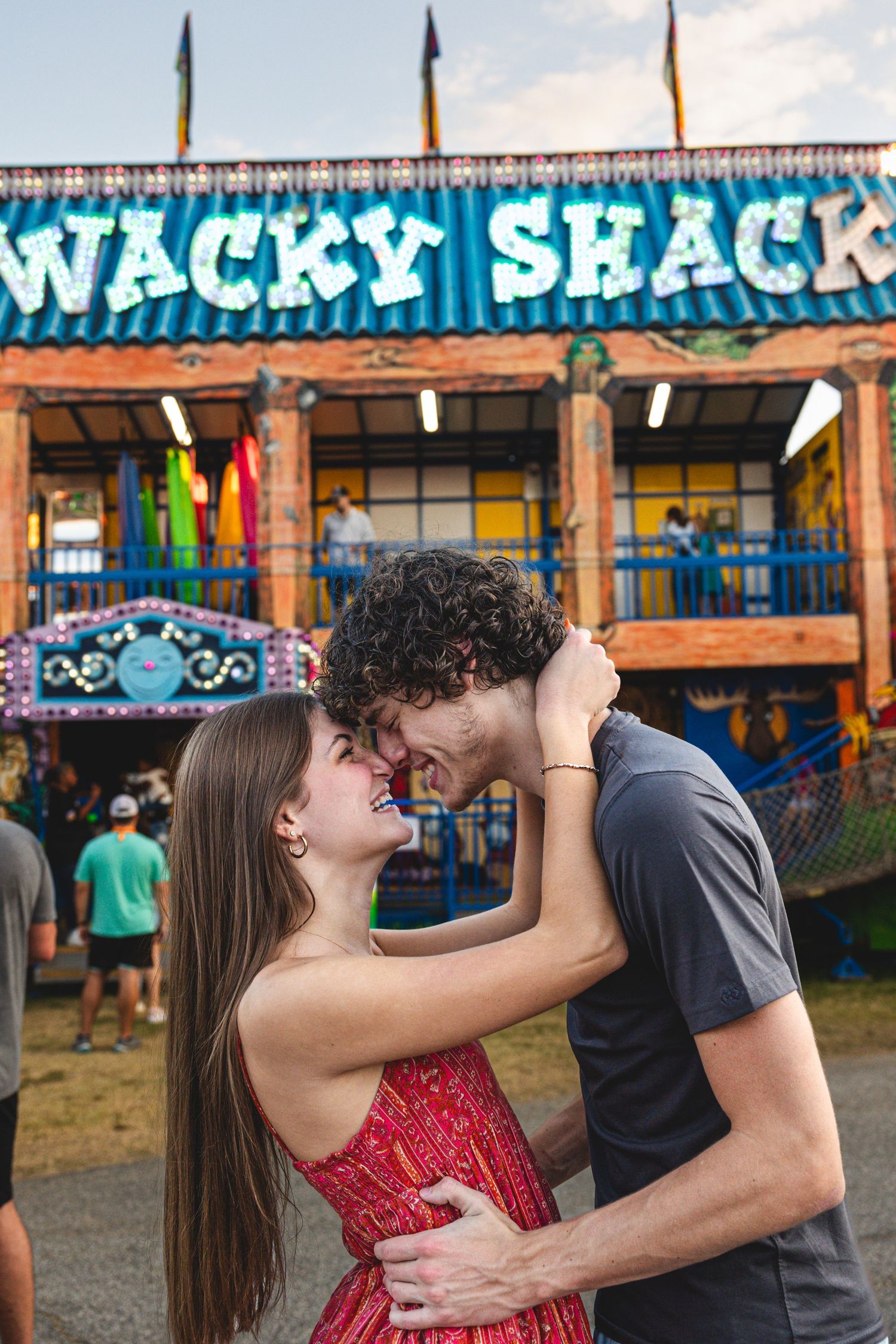 Romantic couple embracing at a carnival in front of a colorful Wacky Shack attraction.