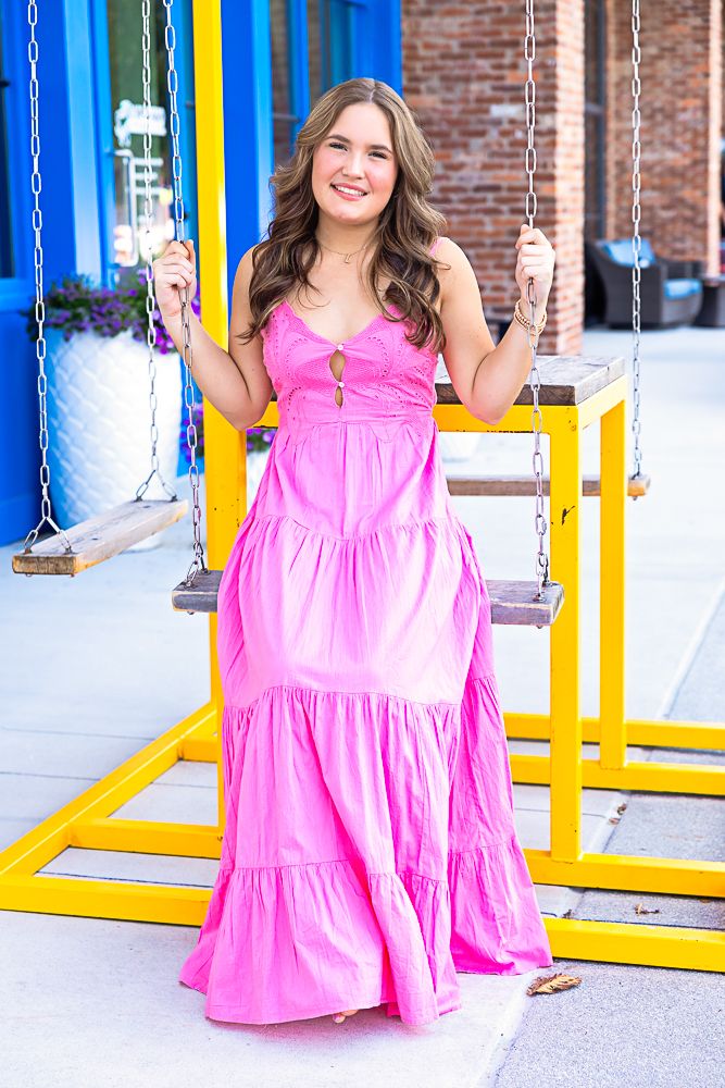 Woman in bright pink tiered dress sits on playground swing set against brick wall background.