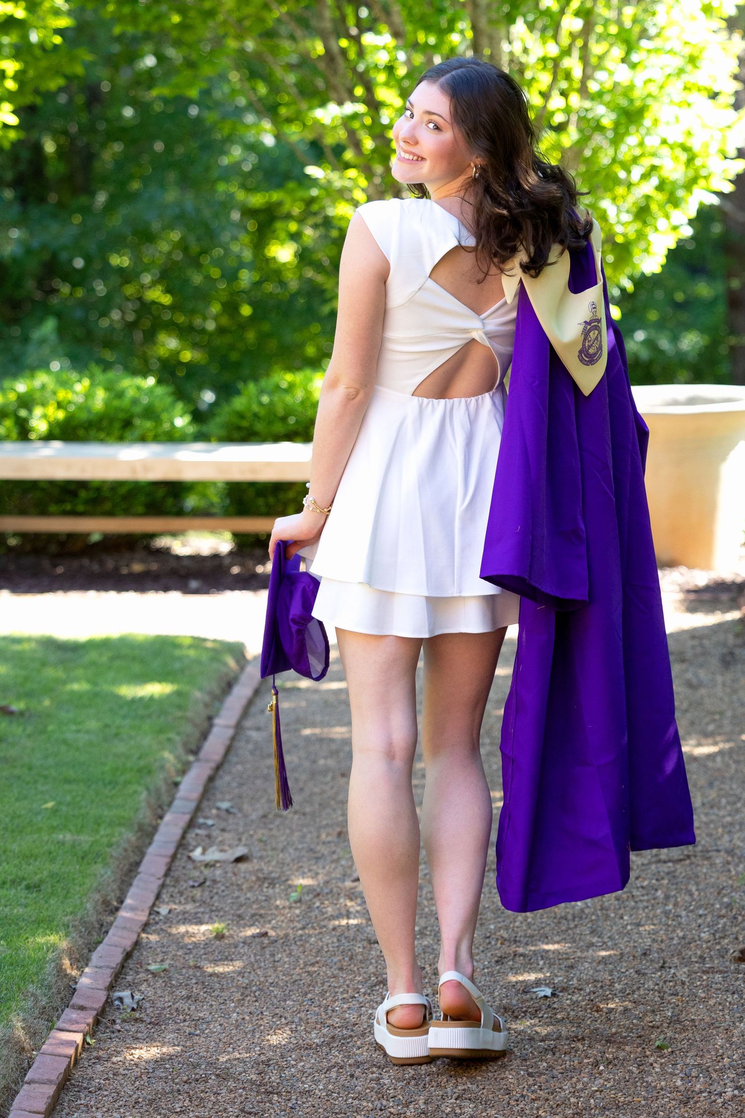 Young woman in white dress holding purple graduation gown and cap, posing outdoors on a brick path surrounded by greenery.