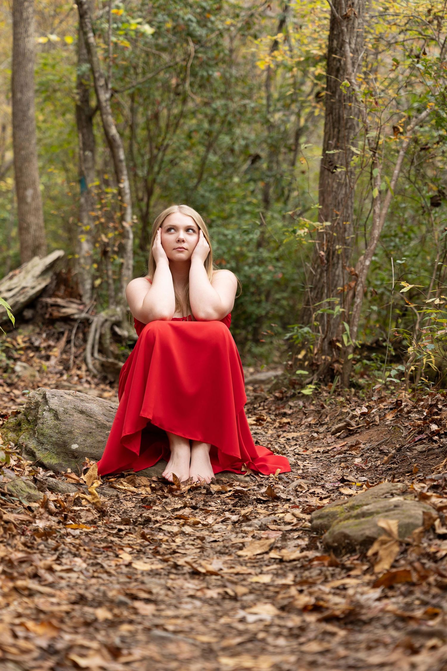 Someone in a flowing red dress sits on a forest path covered in autumn leaves while looking upward.