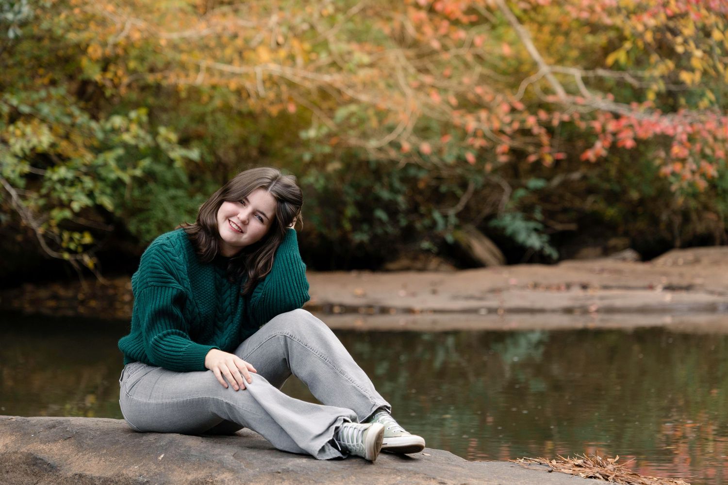 Person in casual attire relaxes by a tranquil pond surrounded by autumn foliage in a peaceful park setting.
