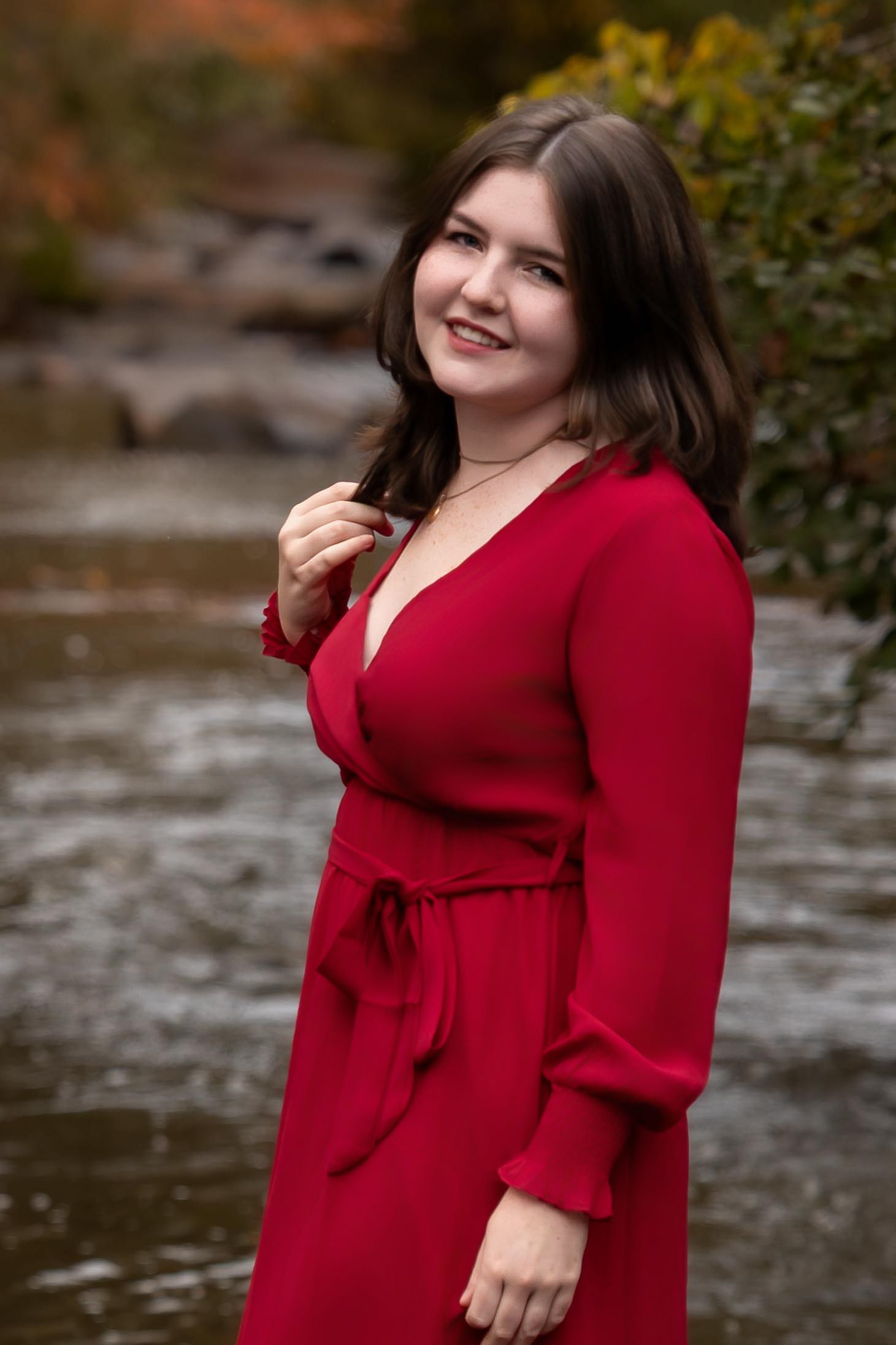 A person in a red wrap dress poses outdoors near water during autumn with trees in the background.