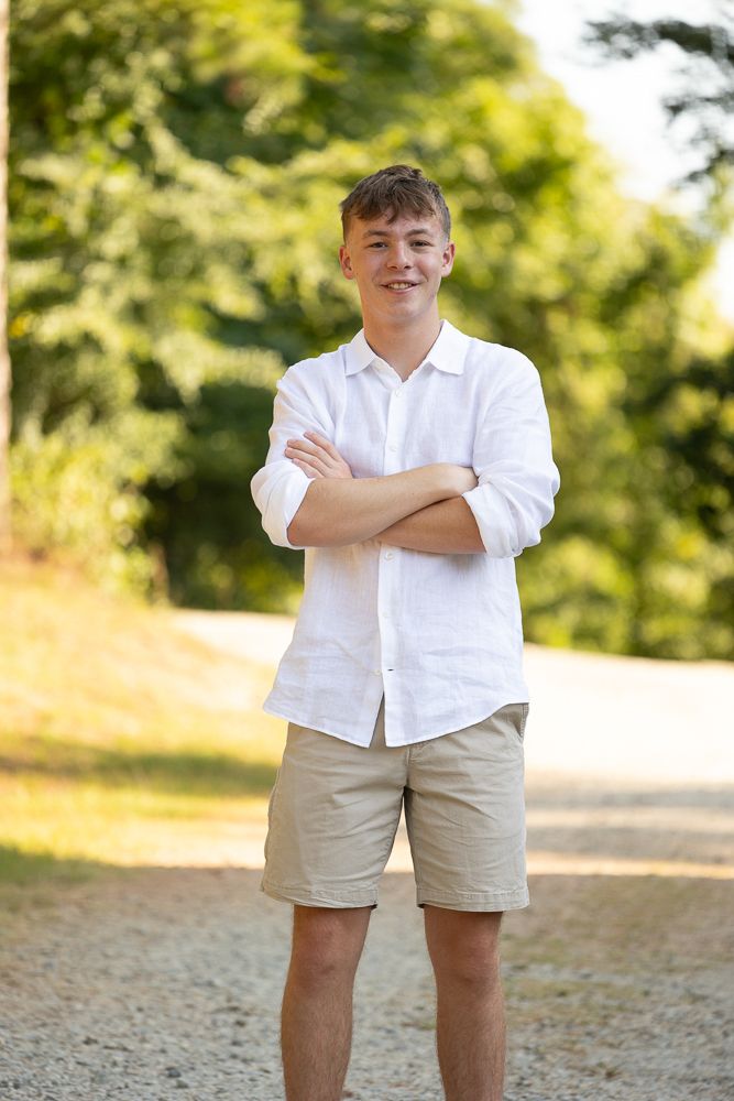 Young man in white linen shirt and khaki shorts stands with arms crossed, smiling outdoors on a sunny day.