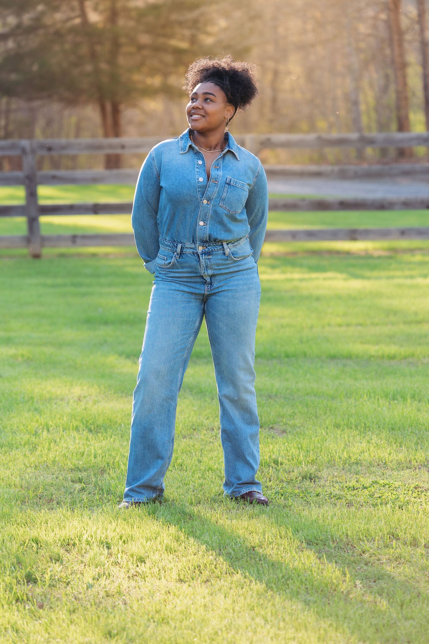 A young Black woman in a denim shirt and jeans stands smiling on green grass near a wooden fence.