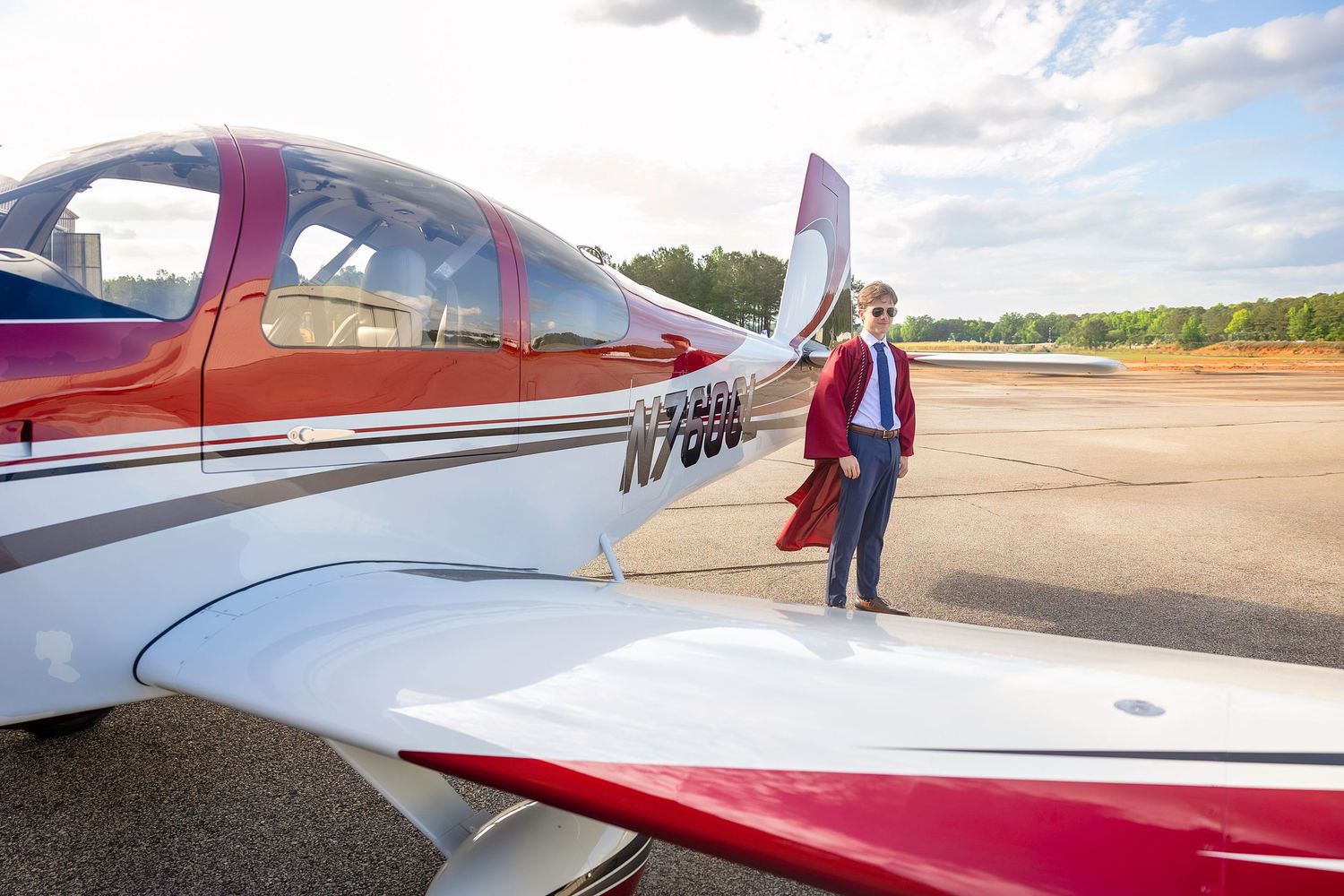 A pilot stands beside a red and white small aircraft on a tarmac, posing confidently near the wing.