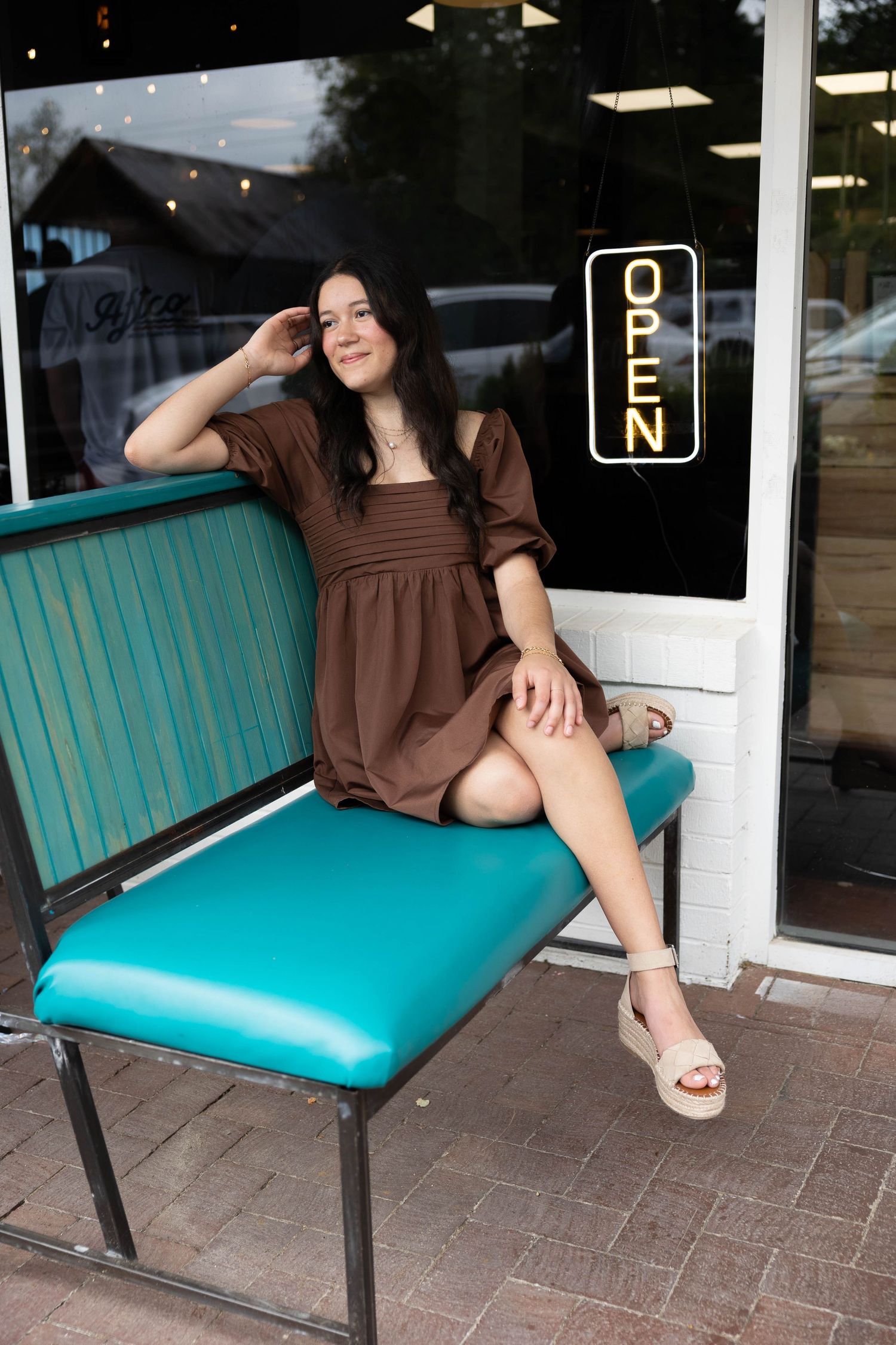A woman in a brown dress sits and poses on a retro turquoise vinyl bench outside a storefront.