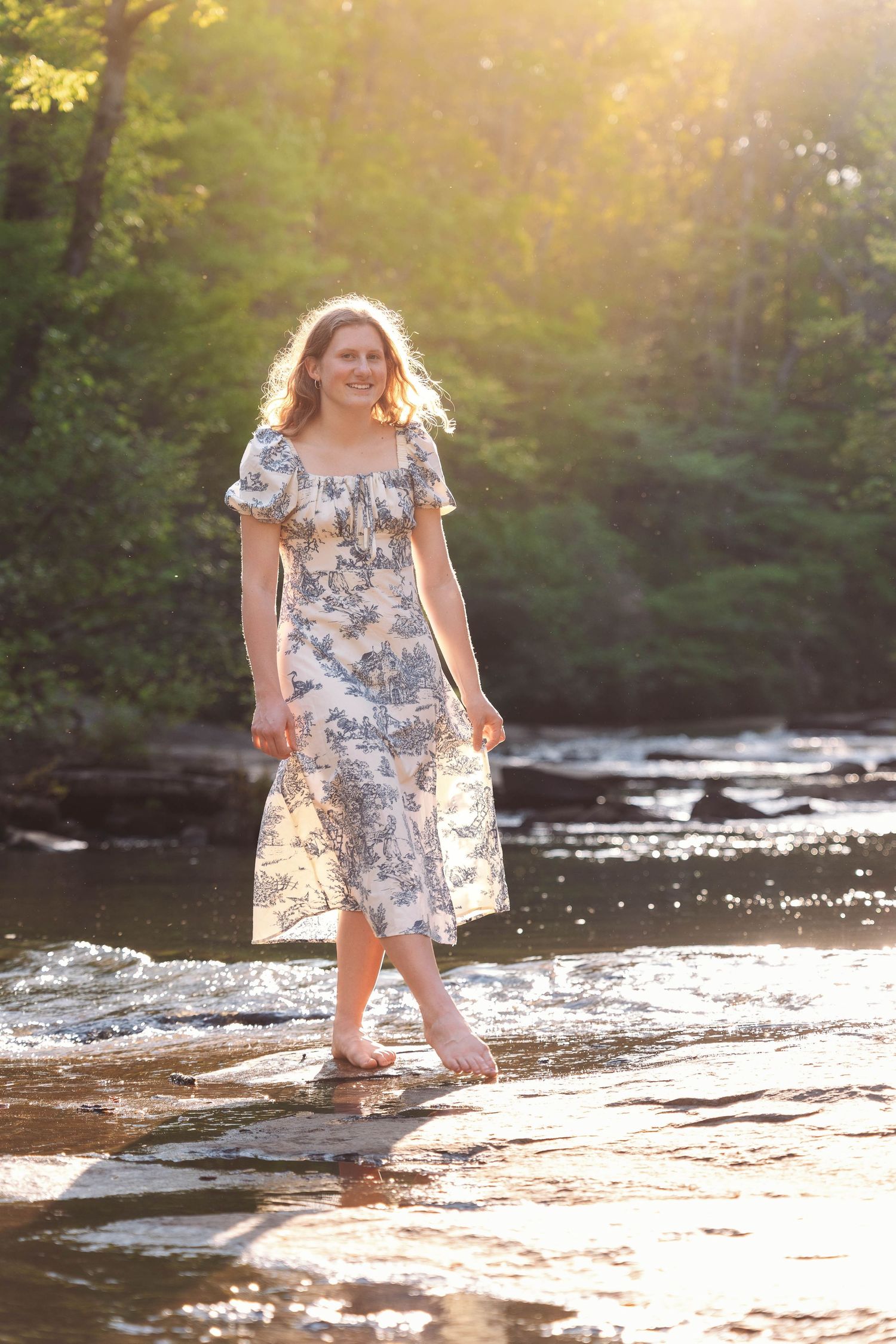 Person in a floral dress stands in shallow river water during golden hour sunlight in a wooded setting.