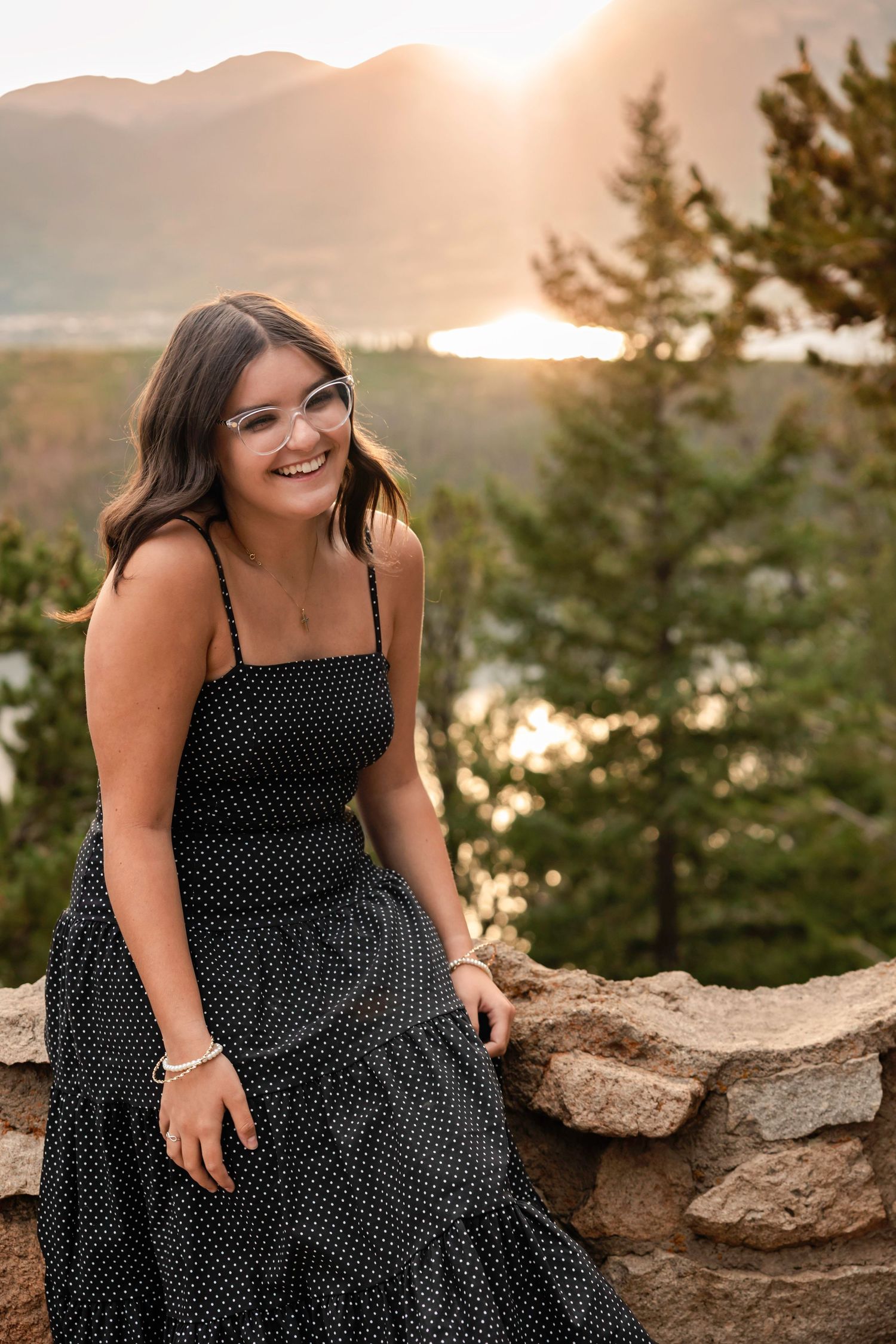 A person in a polka dot dress sits on rocky cliffs overlooking a lake at sunset.