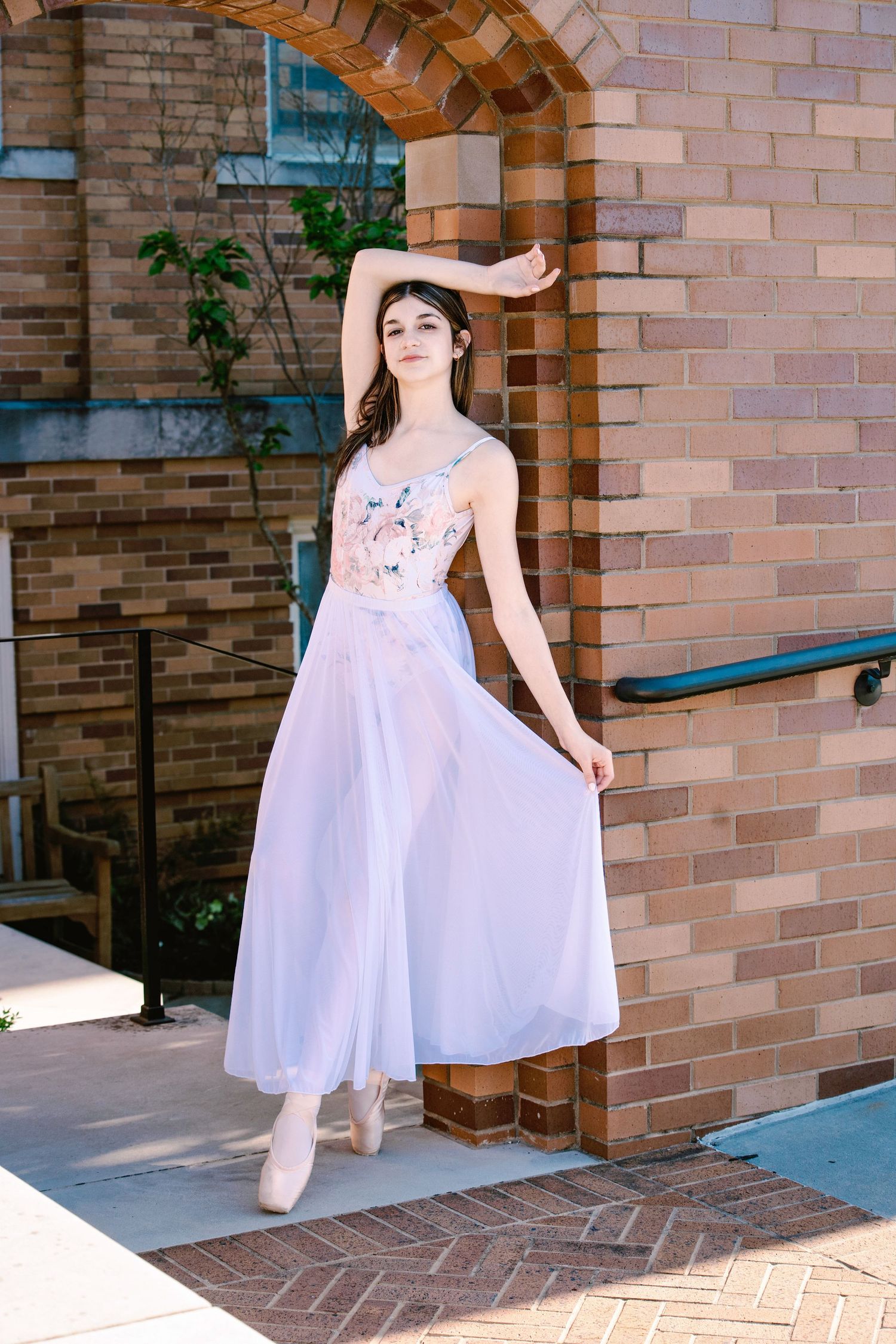 A graceful figure in a flowing white dress and ballet shoes poses elegantly beneath a brick archway.