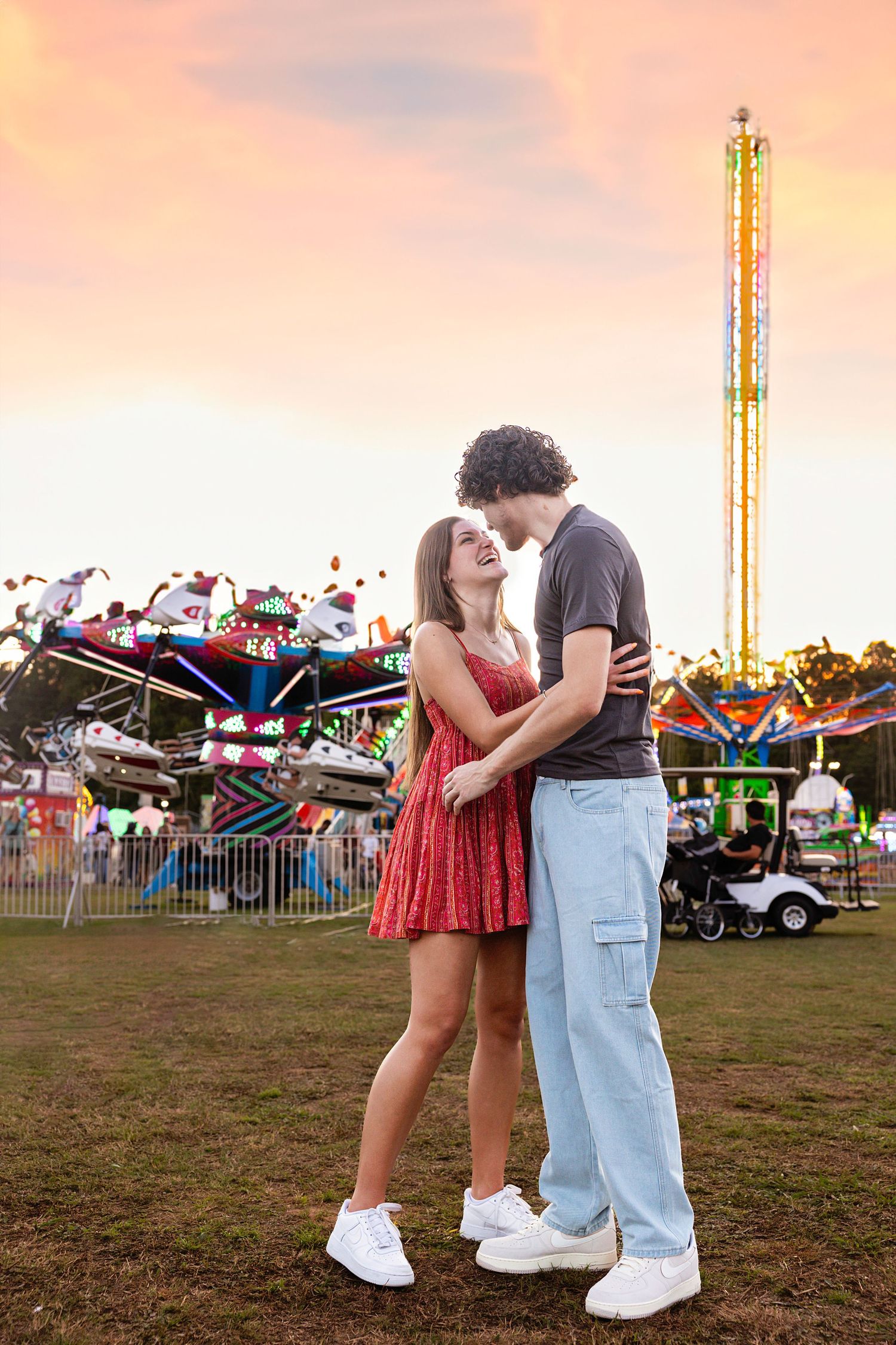 A romantic couple sharing a sweet moment at a carnival with colorful rides in the background.