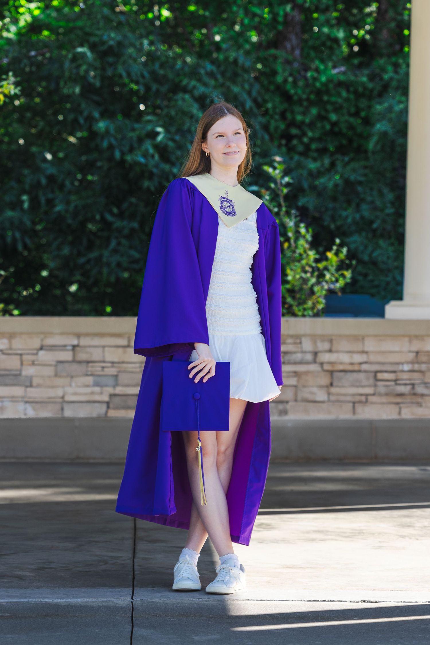 Young woman in purple graduation gown and white dress posing outdoors at graduation ceremony.