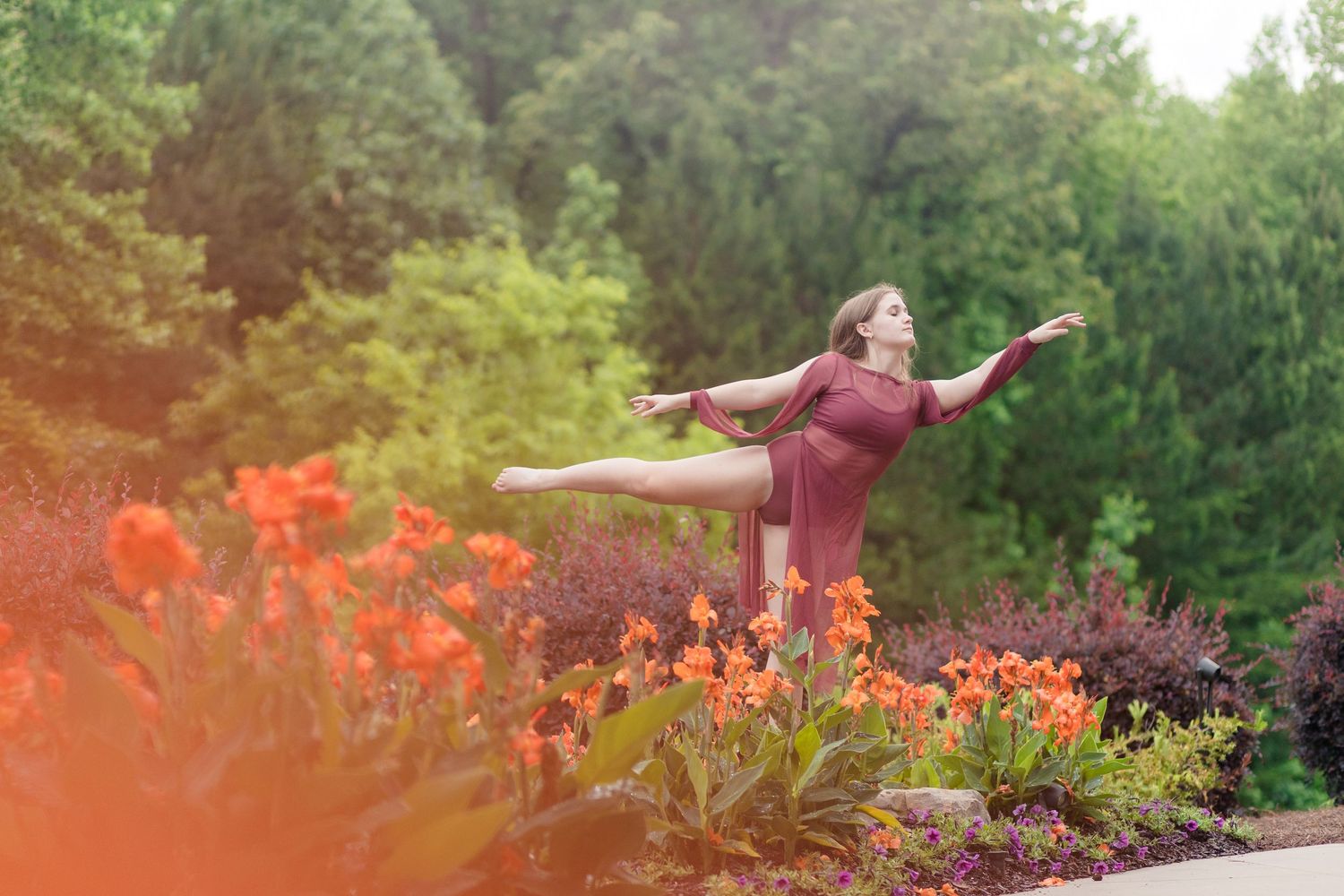 Woman performing graceful ballet pose in lush garden with vibrant orange flowers and green trees in background.
