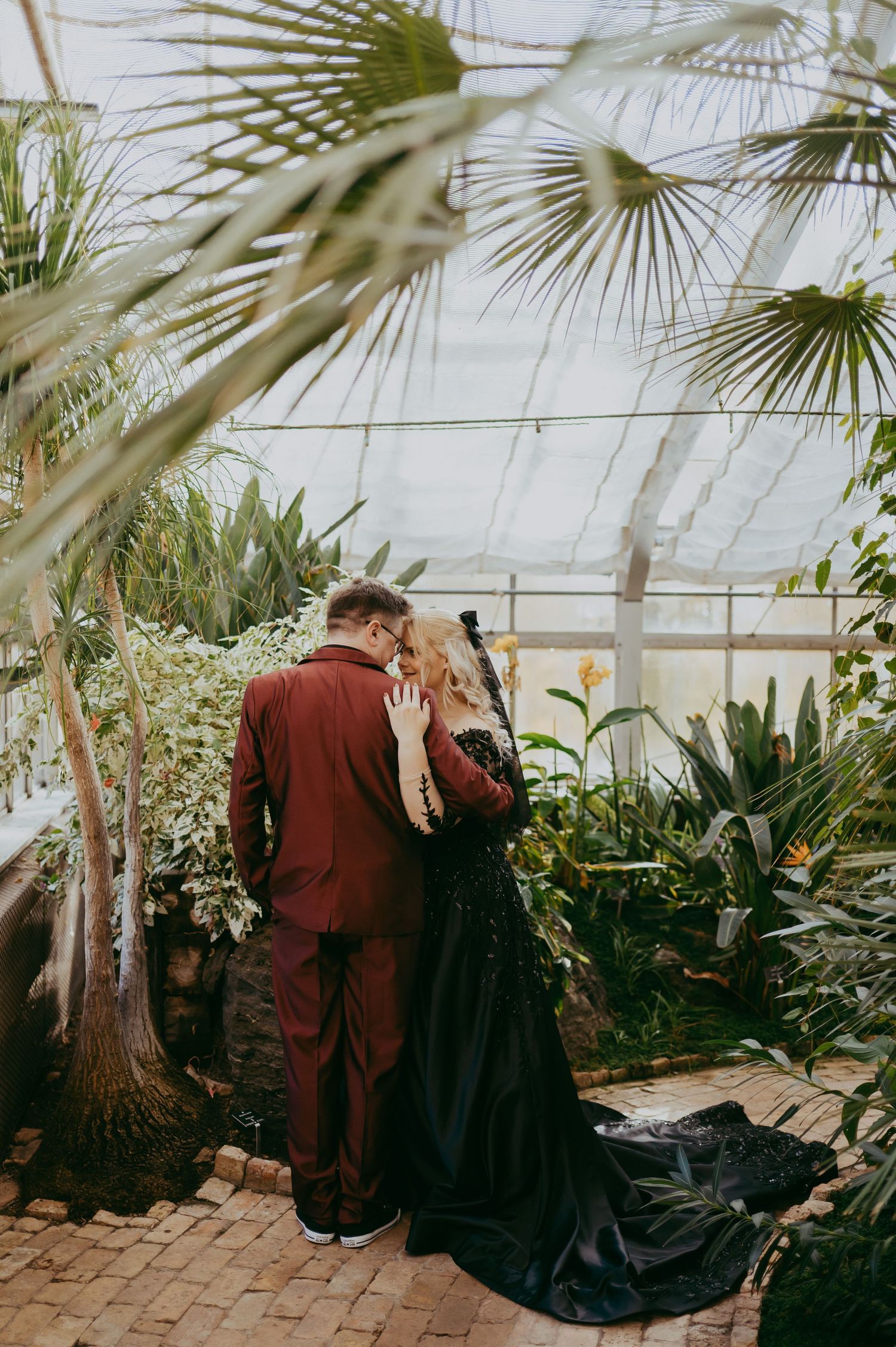 A couple embraces in a greenhouse surrounded by tropical plants, palm fronds and brick flooring.