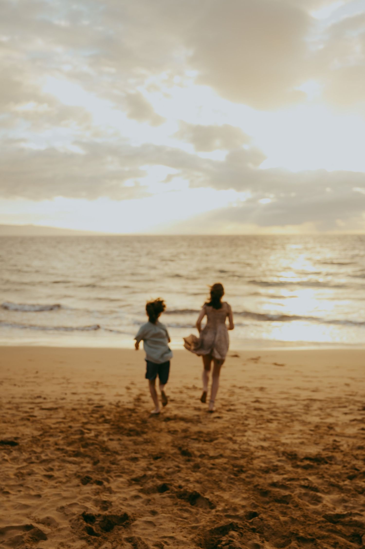 Two people run playfully towards the ocean on a sandy beach at sunset with warm golden light.