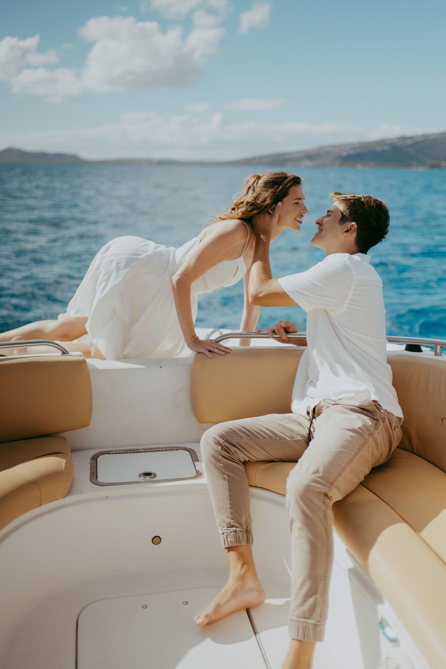 Couple in white outfits share romantic moments on a luxury boat against blue ocean waters and cloudy sky.