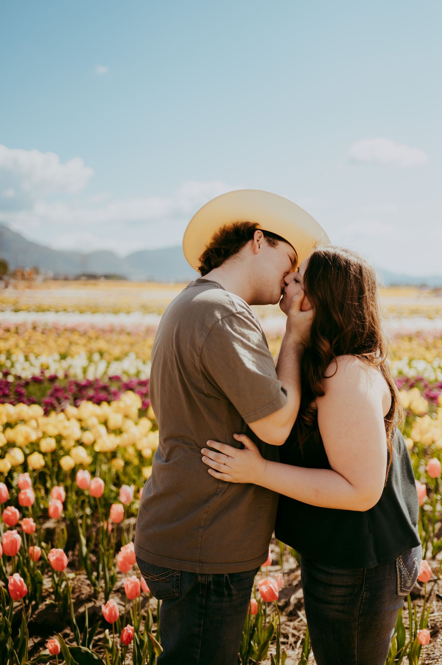 A couple shares a romantic embrace in a vibrant tulip field with mountains and blue sky in the background.