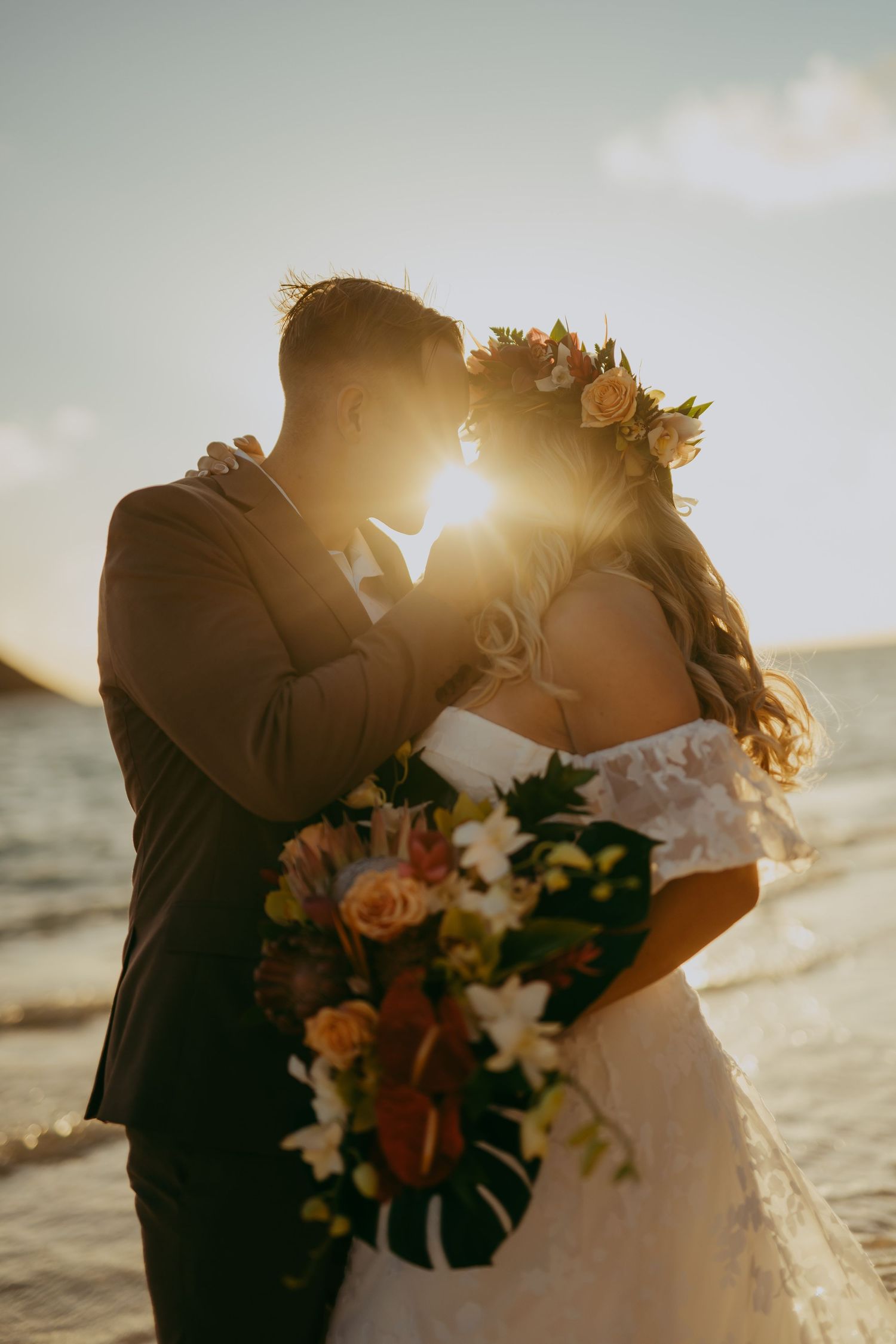 A romantic beach wedding couple embraces during golden hour sunset with tropical flowers and ocean waves in background.