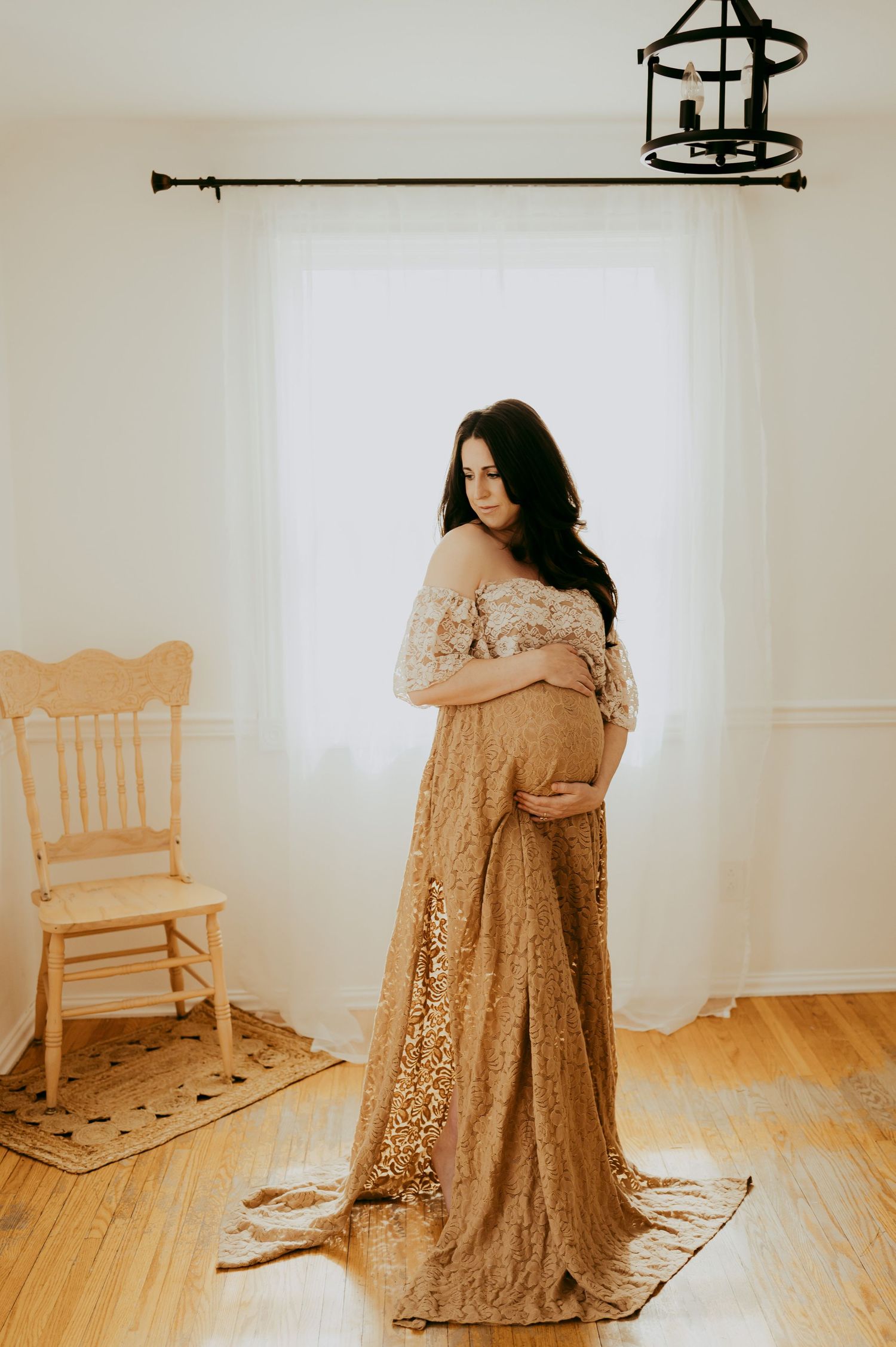 A maternity photo featuring a flowing beige lace gown in a minimalist room with a wooden chair and hardwood floors.