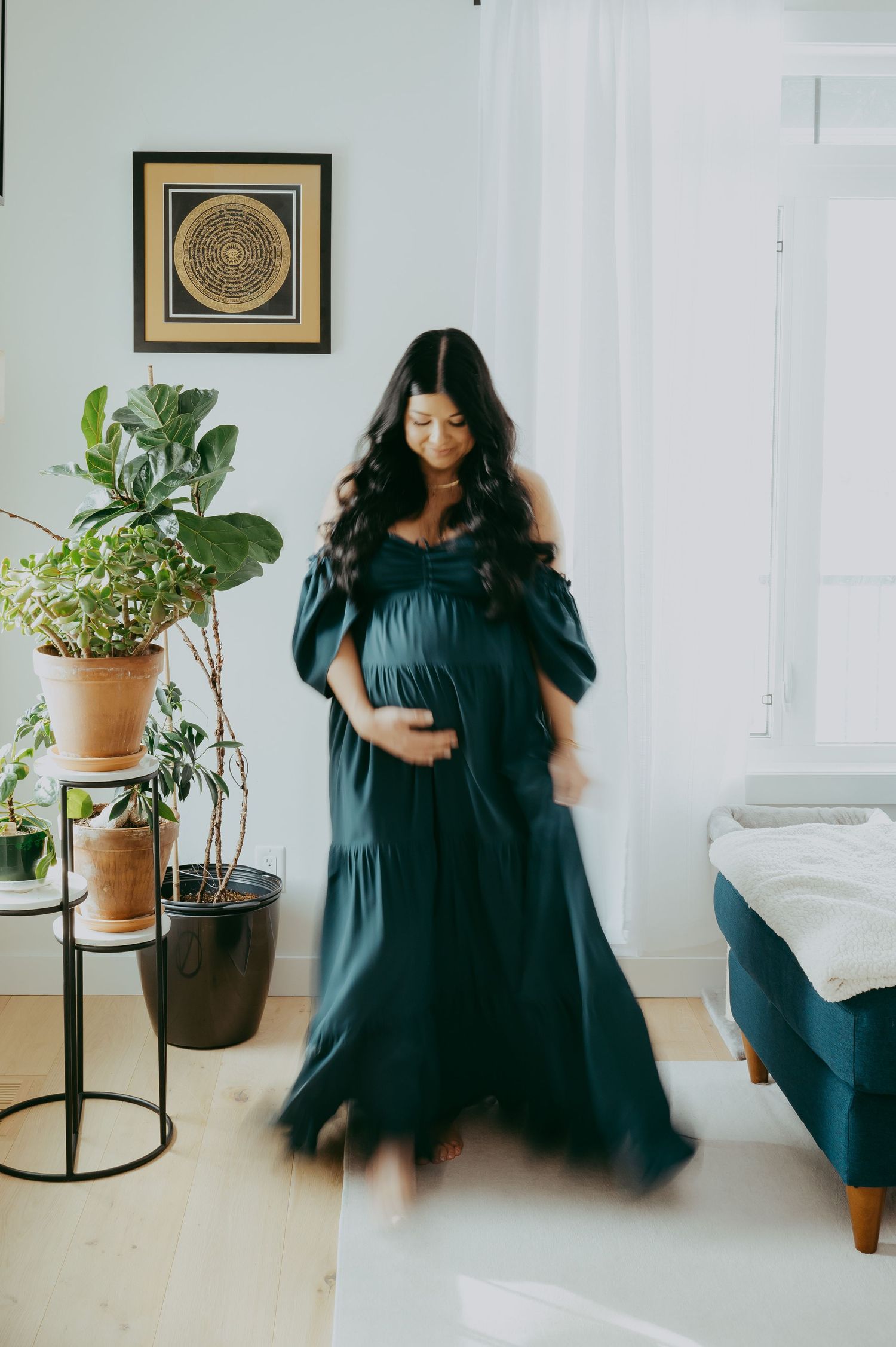 A pregnant person in a flowing navy blue dress stands in a minimalist room with potted plants and framed wall art.