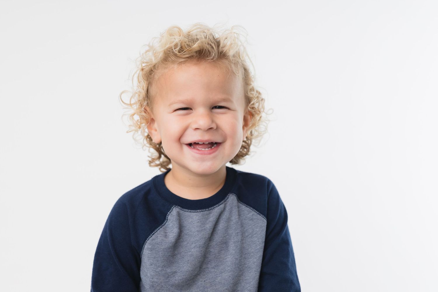 A happy child with curly blonde hair wearing a navy and gray raglan shirt against a white background.