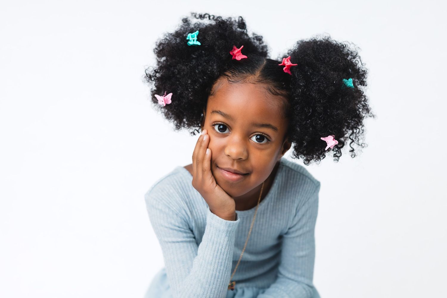 A young child wearing a light gray sweater rests their chin in their hand while smiling at the camera against a white background.