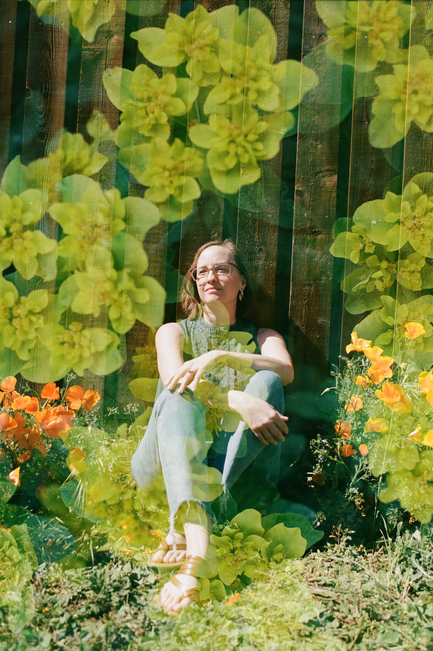 Person sits in casual summer attire against wooden fence with yellow and orange flowers blooming around them.