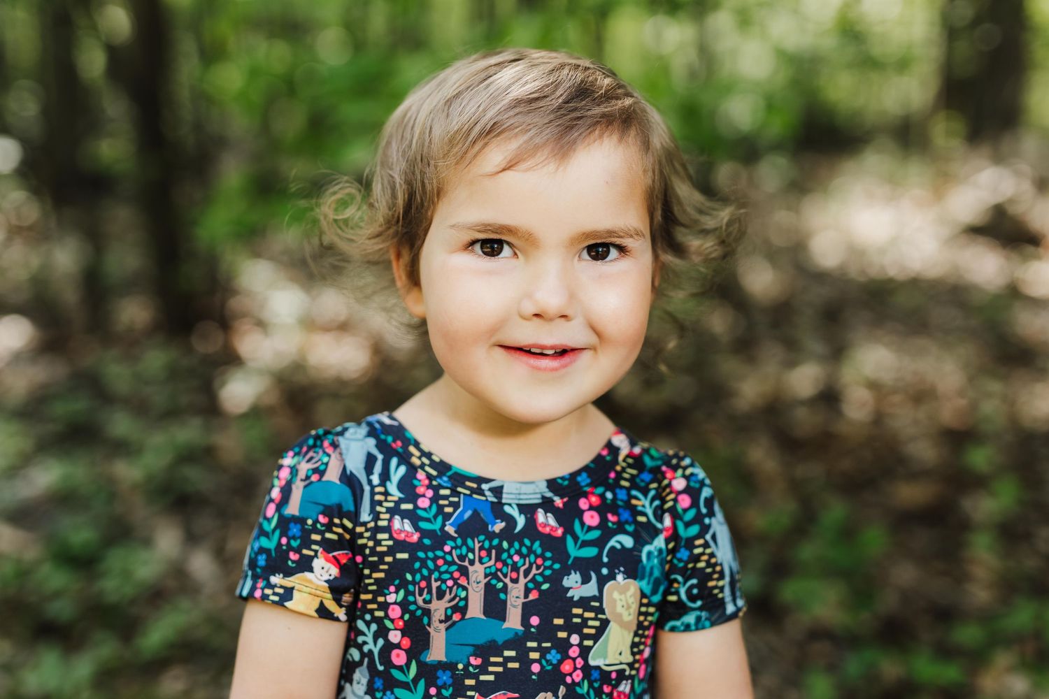 A young child smiles while wearing a colorful floral dress outdoors in a natural setting.