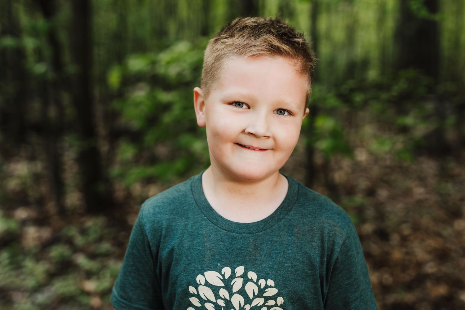 Young person wearing a dark green shirt poses outdoors in a forest setting with natural light and foliage in background.