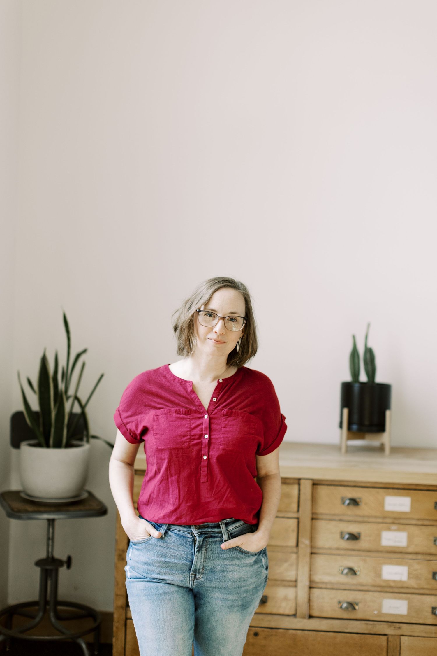Person in casual red top and jeans standing near houseplants and wooden furniture.