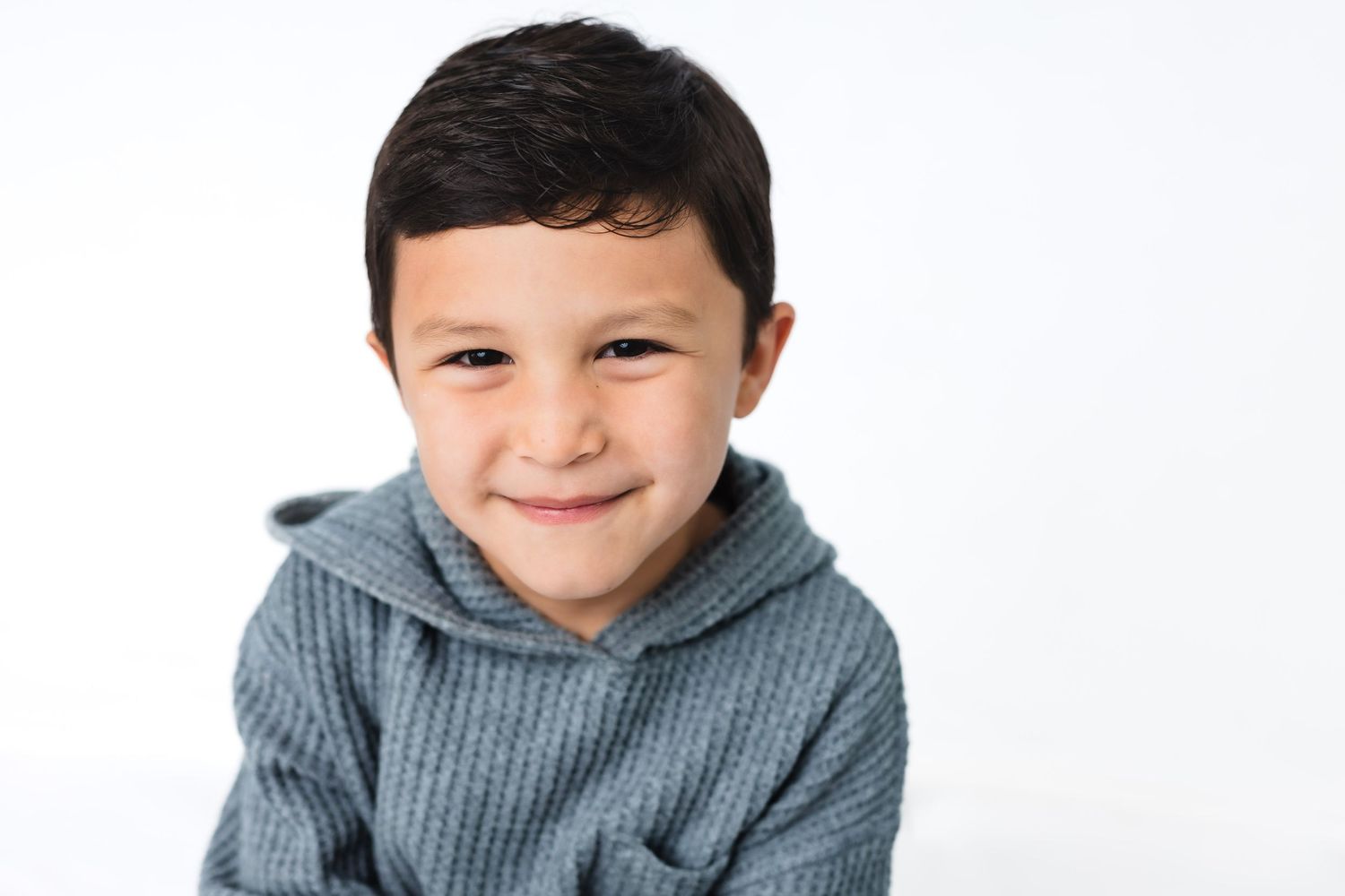 A young child wearing a gray knit sweater smiles while posing for a casual photo series against a white background.