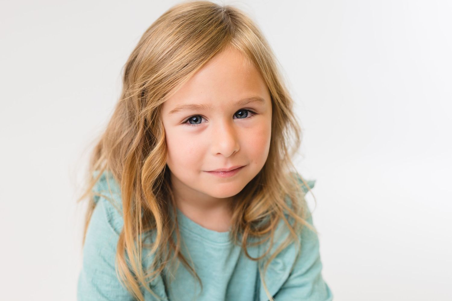 Young child wearing a light blue top smiles brightly while sitting against a white background.